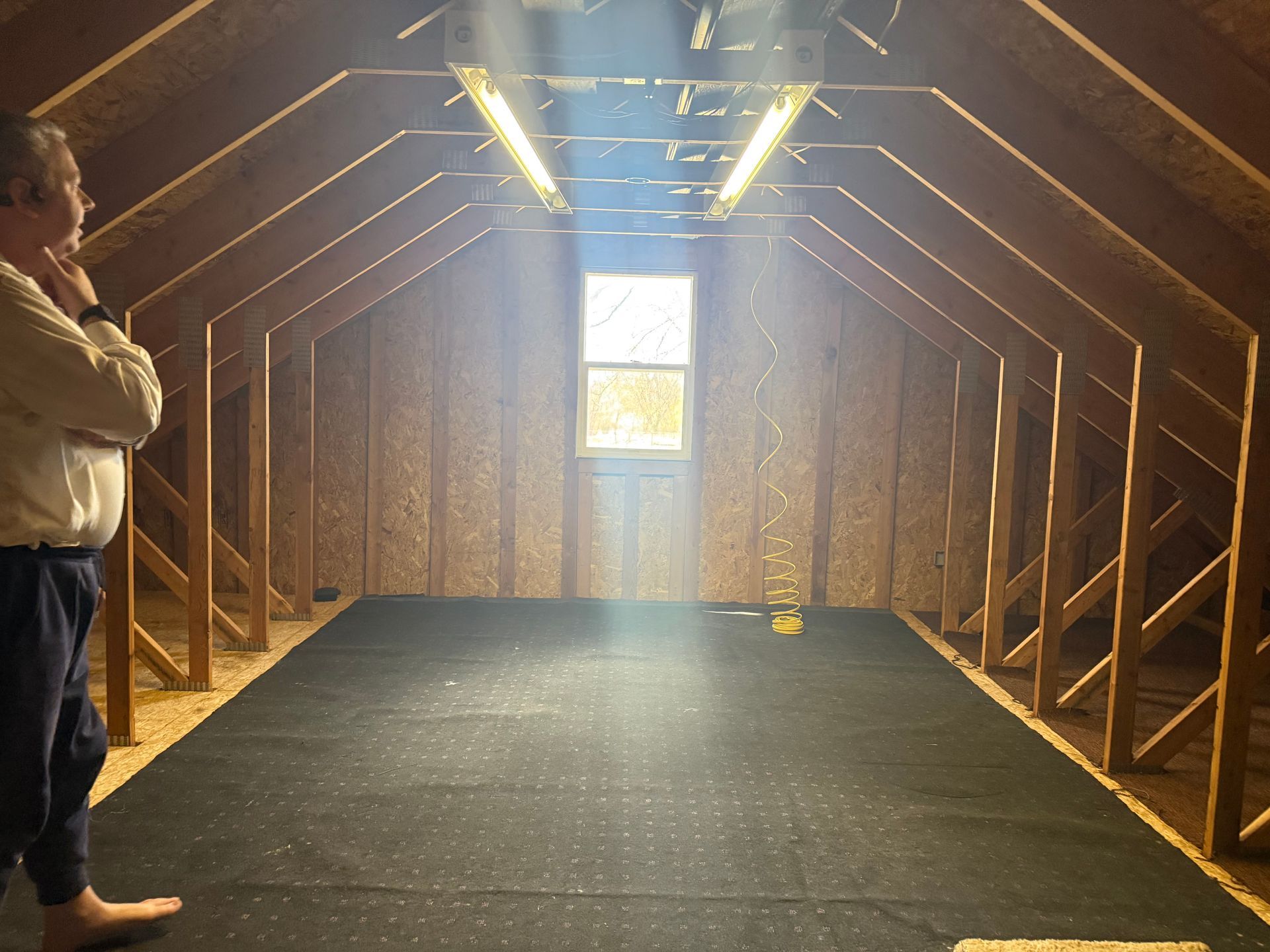 A person stands in a sunlit unfinished attic with wooden rafters, a window, and a dark subfloor.