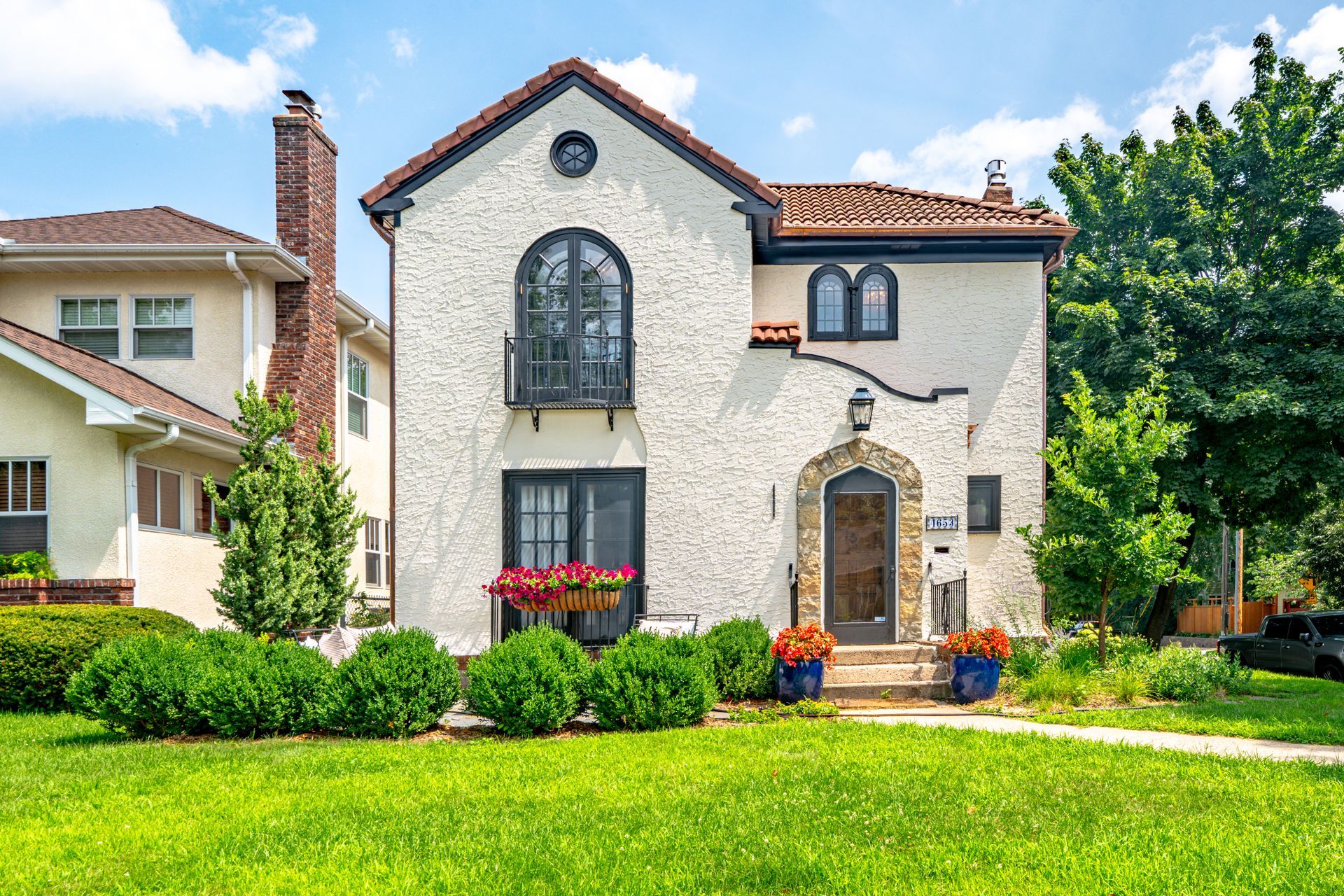 A two-story, white stucco house with a tiled roof, arched window, and small balcony, set behind a lush green lawn.