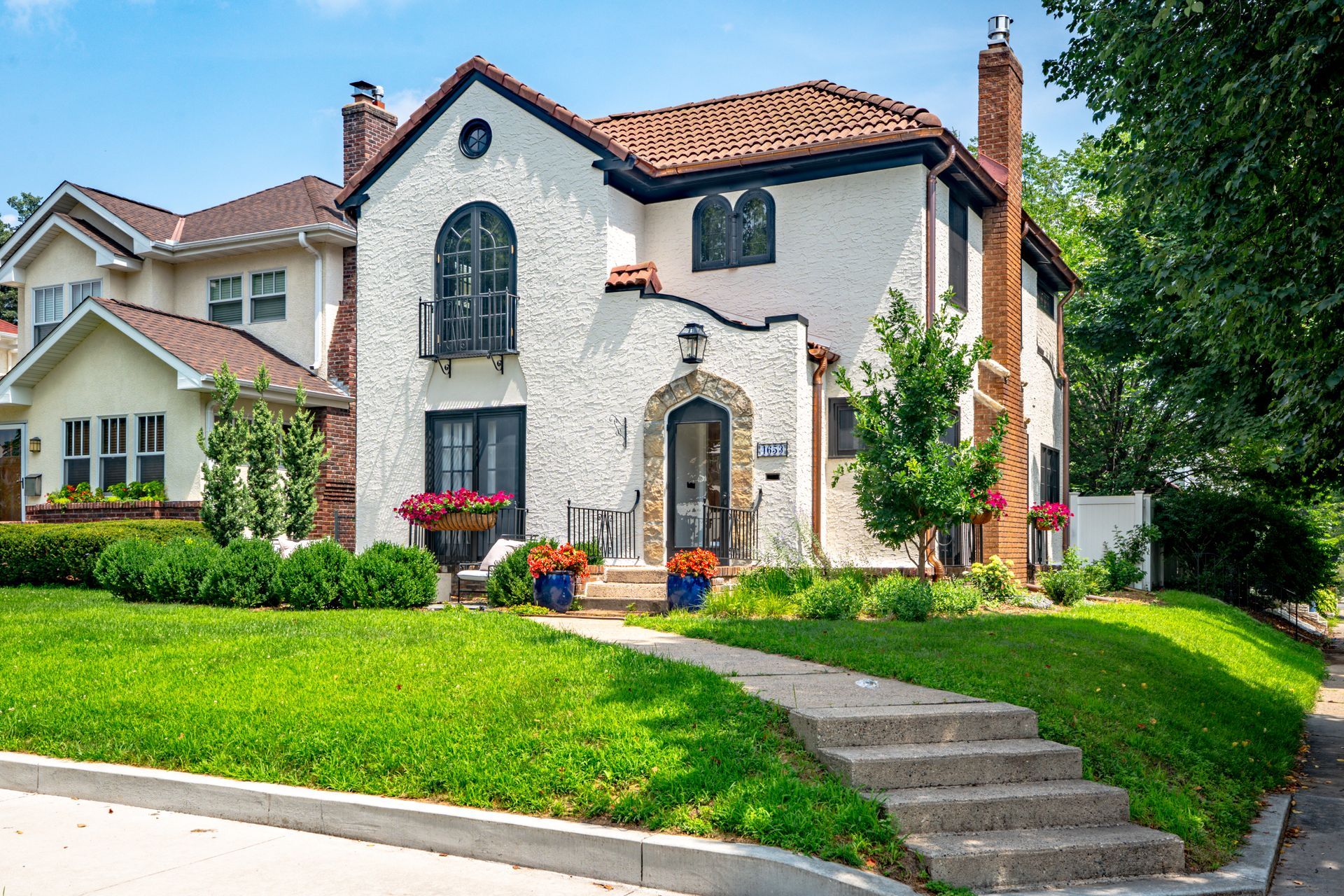 A two-story, white stucco Spanish-style house with a brown tile roof, stone arched entrance, and a green front lawn.