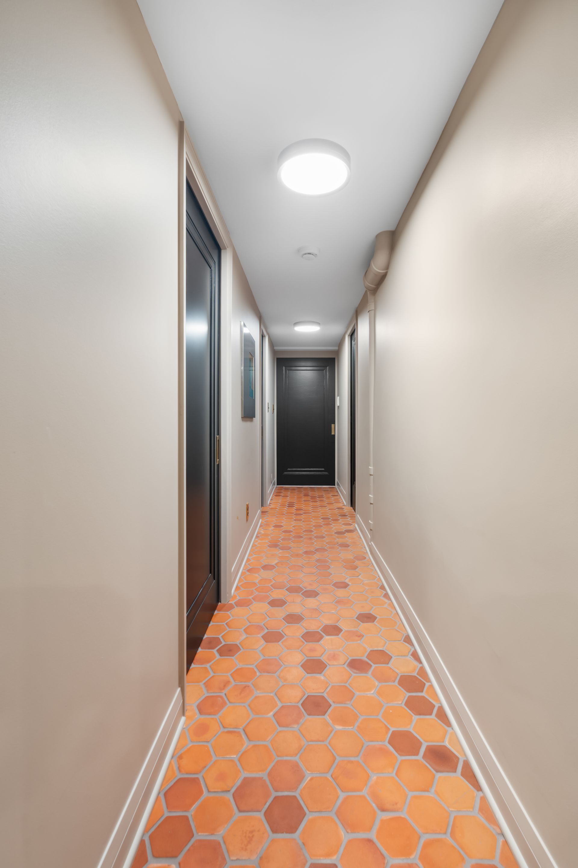 A long, narrow hallway with light tan walls, a white ceiling with two circular lights, and orange hexagonal tile flooring.