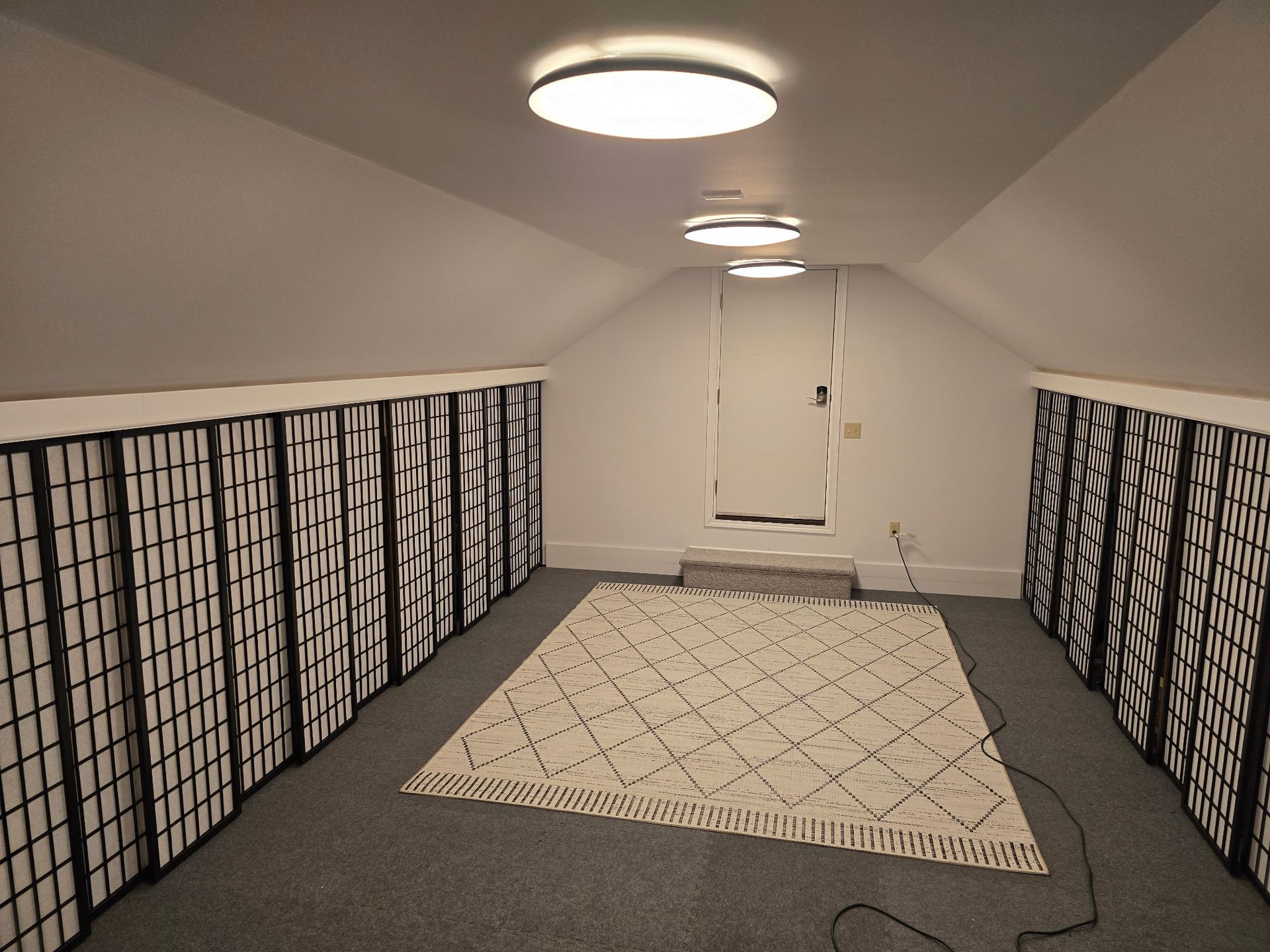 An attic room with a rug on gray carpet, framed by walls lined with black-and-white patterned folding screens.