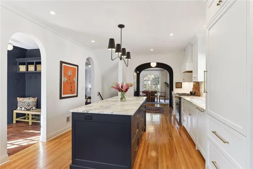 A bright, white kitchen with a dark blue island, marble countertop, wooden floors, and an arched doorway to a mudroom.