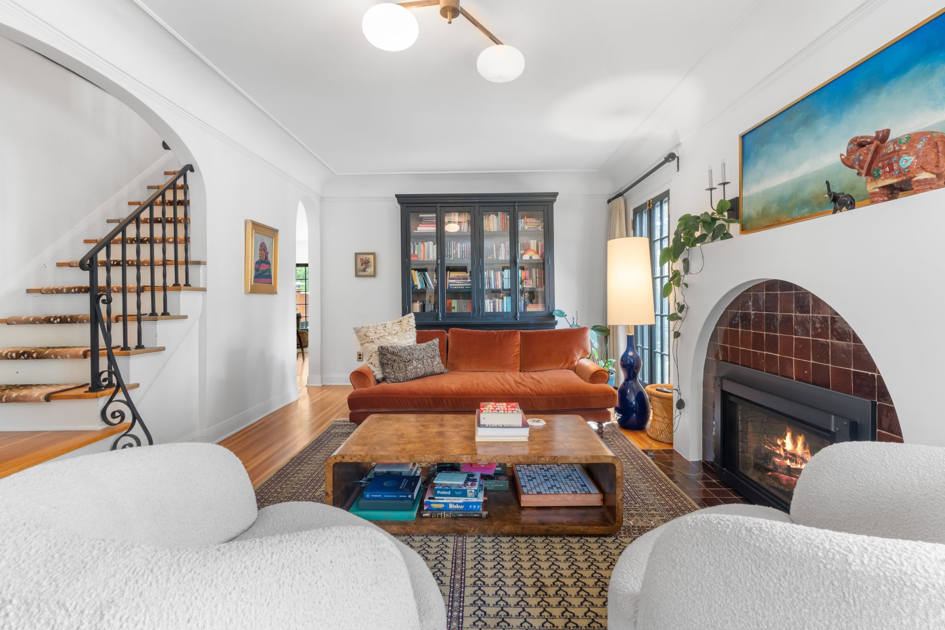 A cozy living room with a white arched fireplace, orange velvet sofa, two white armchairs, and a winding staircase.