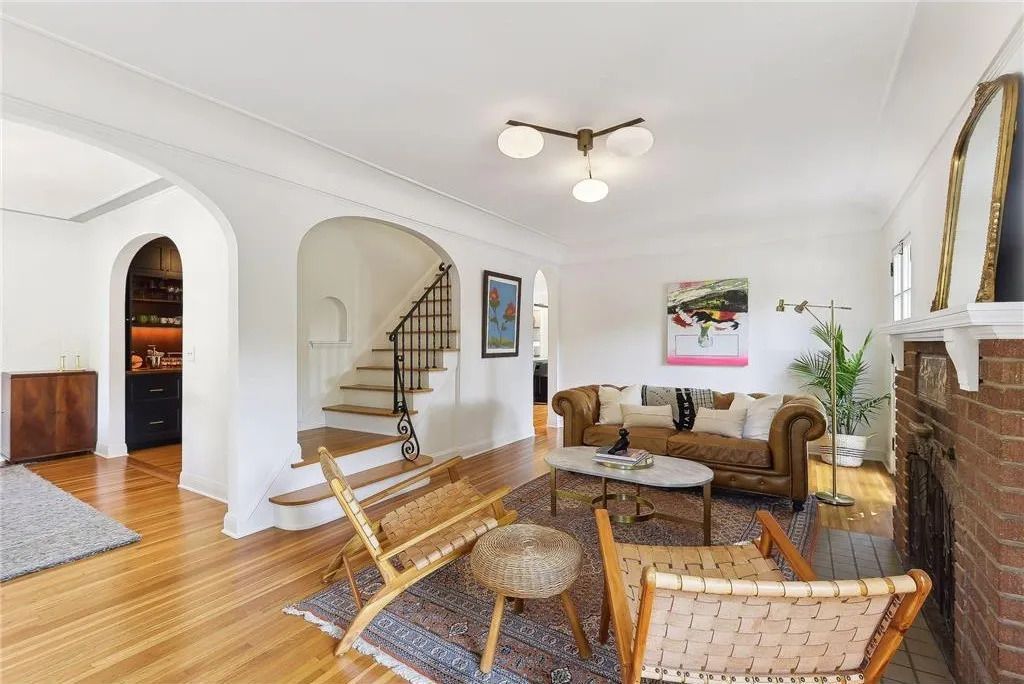A living room with wooden floors, an arched entryway to a staircase, a brown couch, two wood chairs, and a brick fireplace.