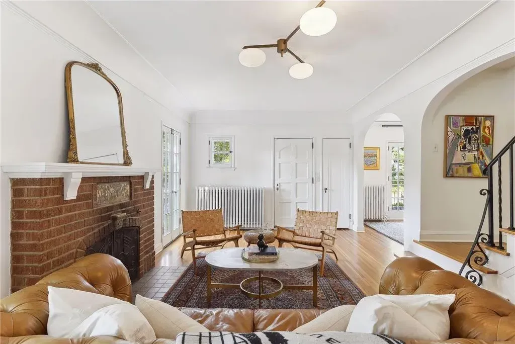 A sunlit living room features a brick fireplace, tan tufted sofa, two wicker chairs, and a central white coffee table.