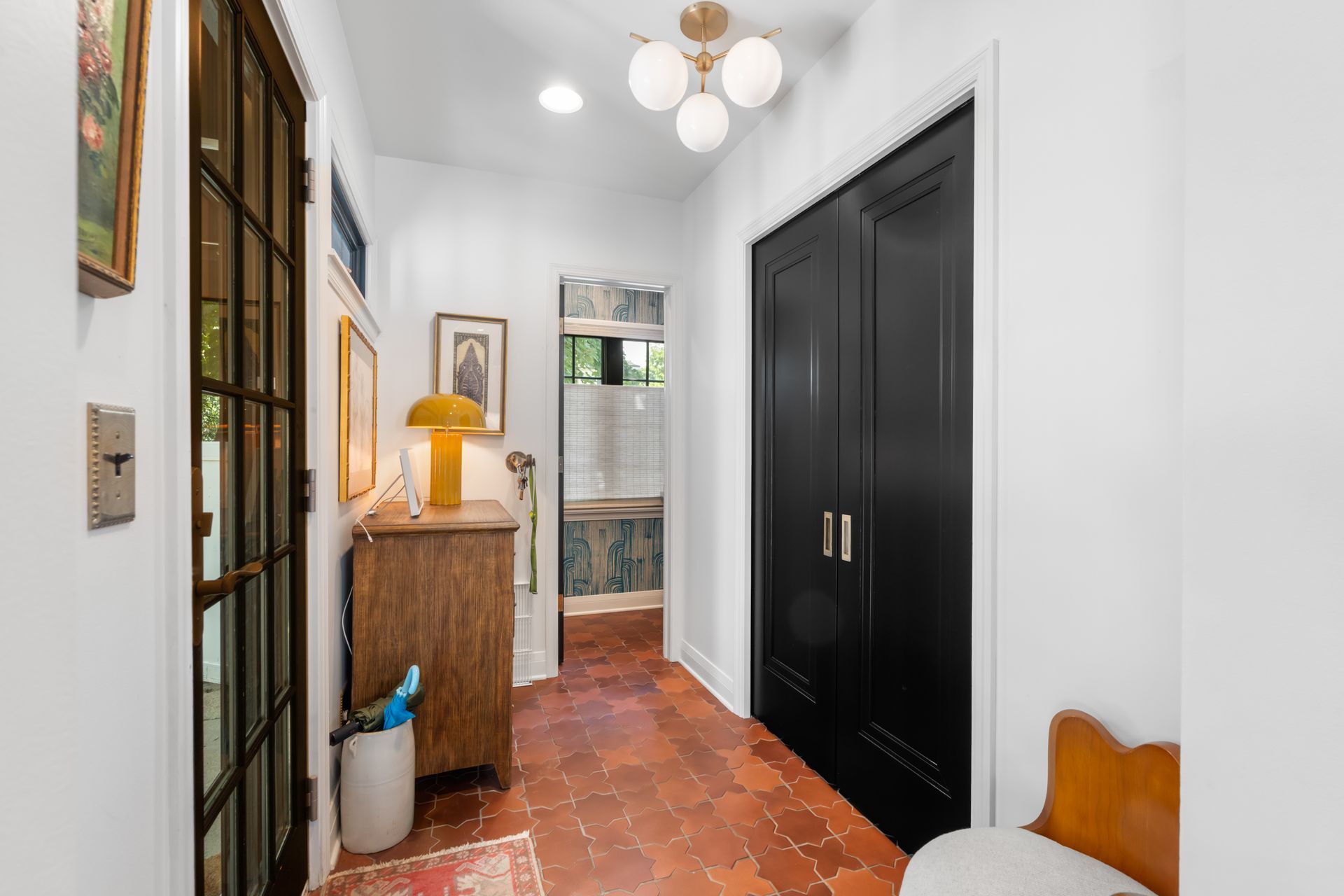 A bright hallway with terracotta tile floors, a wooden cabinet, a modern light fixture, and black double doors.