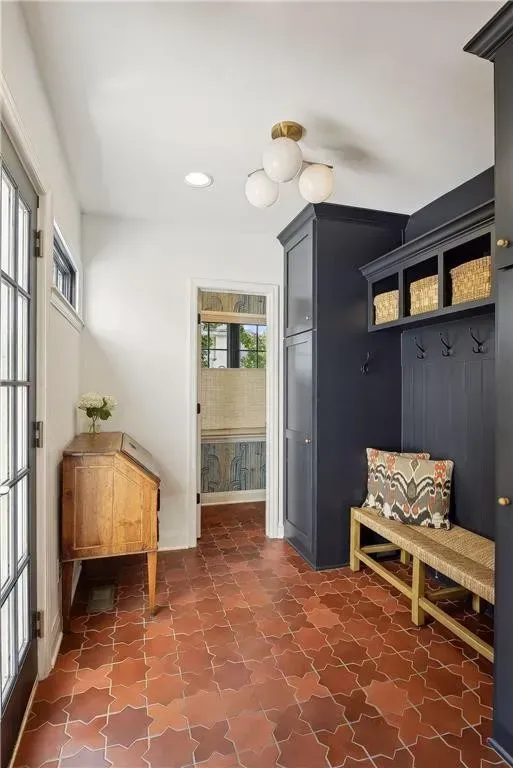 Mudroom with terracotta tiles, a dark blue storage unit with hooks, a wooden bench, and a vintage desk near a glass door.