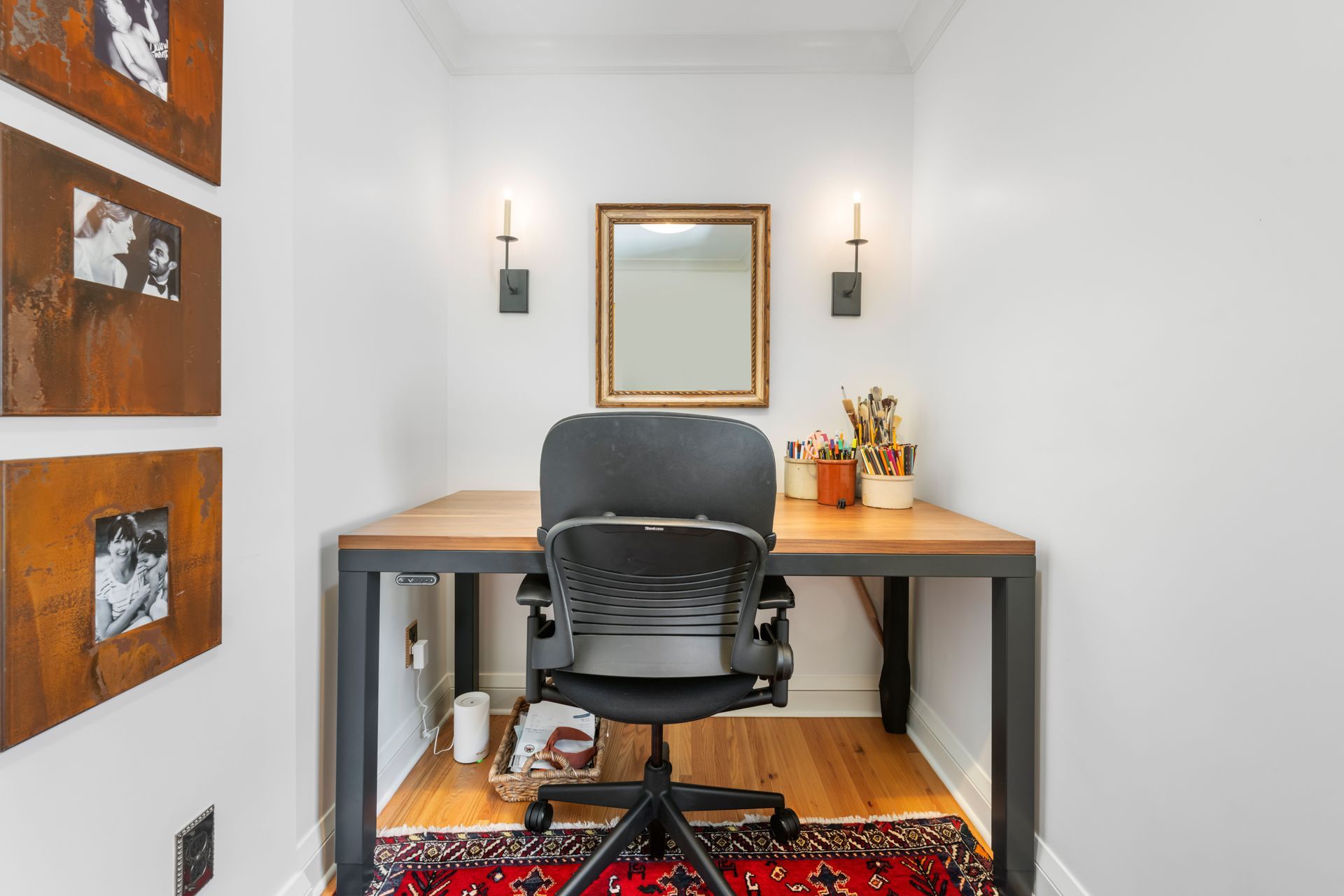 A small home office nook with a desk, black ergonomic chair, framed mirror, wall sconces, and art on the left wall.