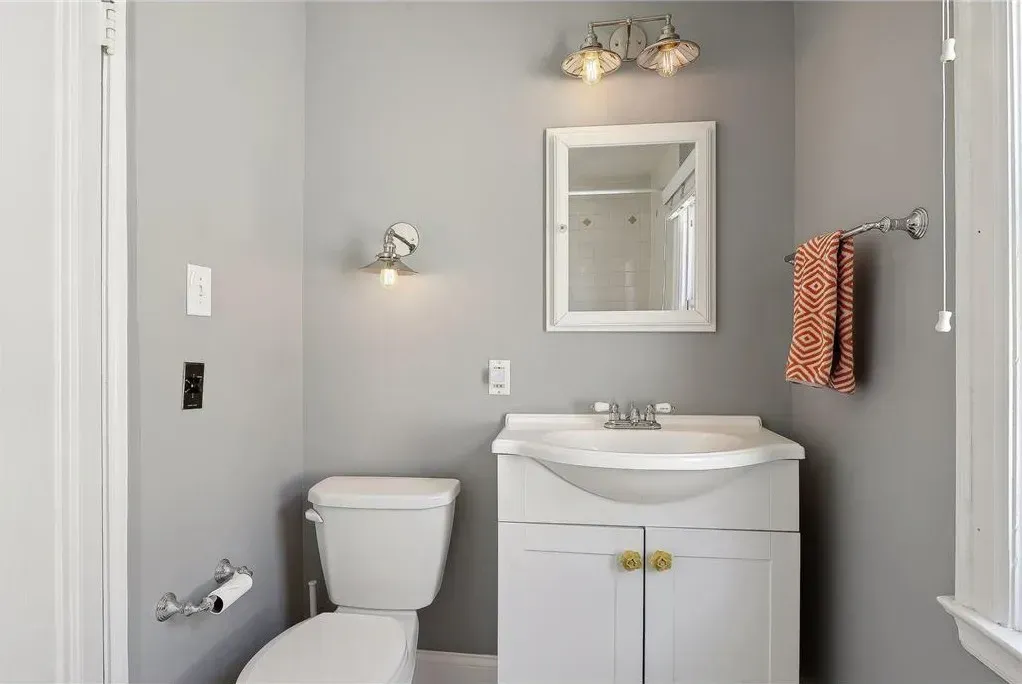 A gray bathroom featuring a white vanity with a pedestal sink, a wall mirror, a toilet, and an orange patterned hand towel.