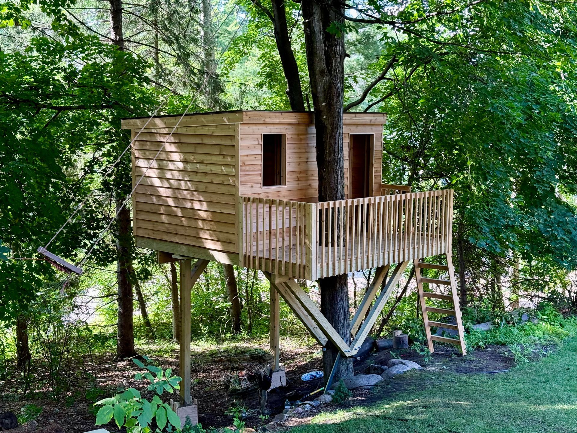 A wooden treehouse with a porch and a ladder, supported by posts and a tree, set in a sunny, forested area.