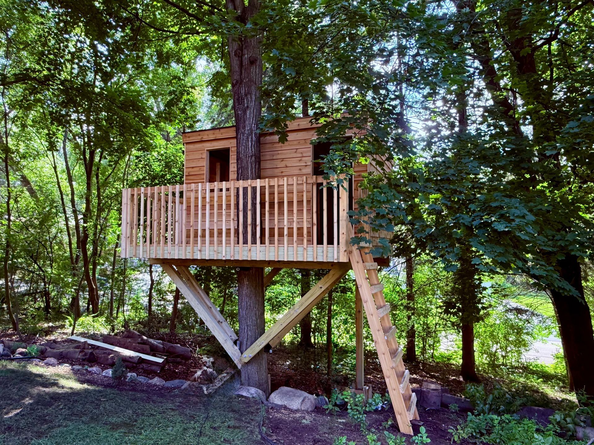 A light-wood treehouse with a balcony and ladder built into a large tree in a lush, wooded area.