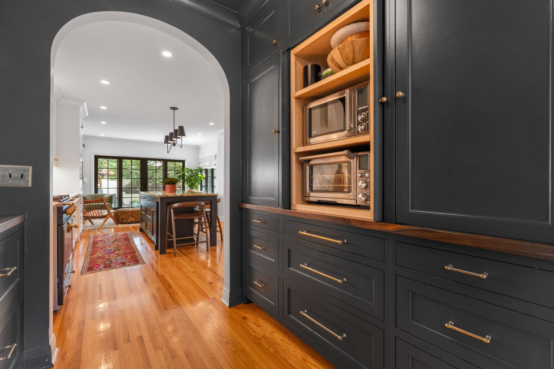 A dark grey kitchen wall with built-in cabinetry and a microwave opens into a bright dining area with a wooden island.