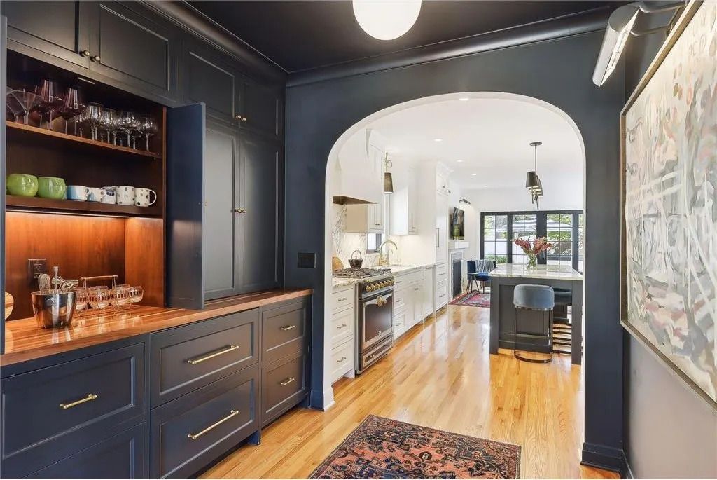 A dark-walled butler's pantry with wood countertops opens into a white kitchen via an arched doorway.