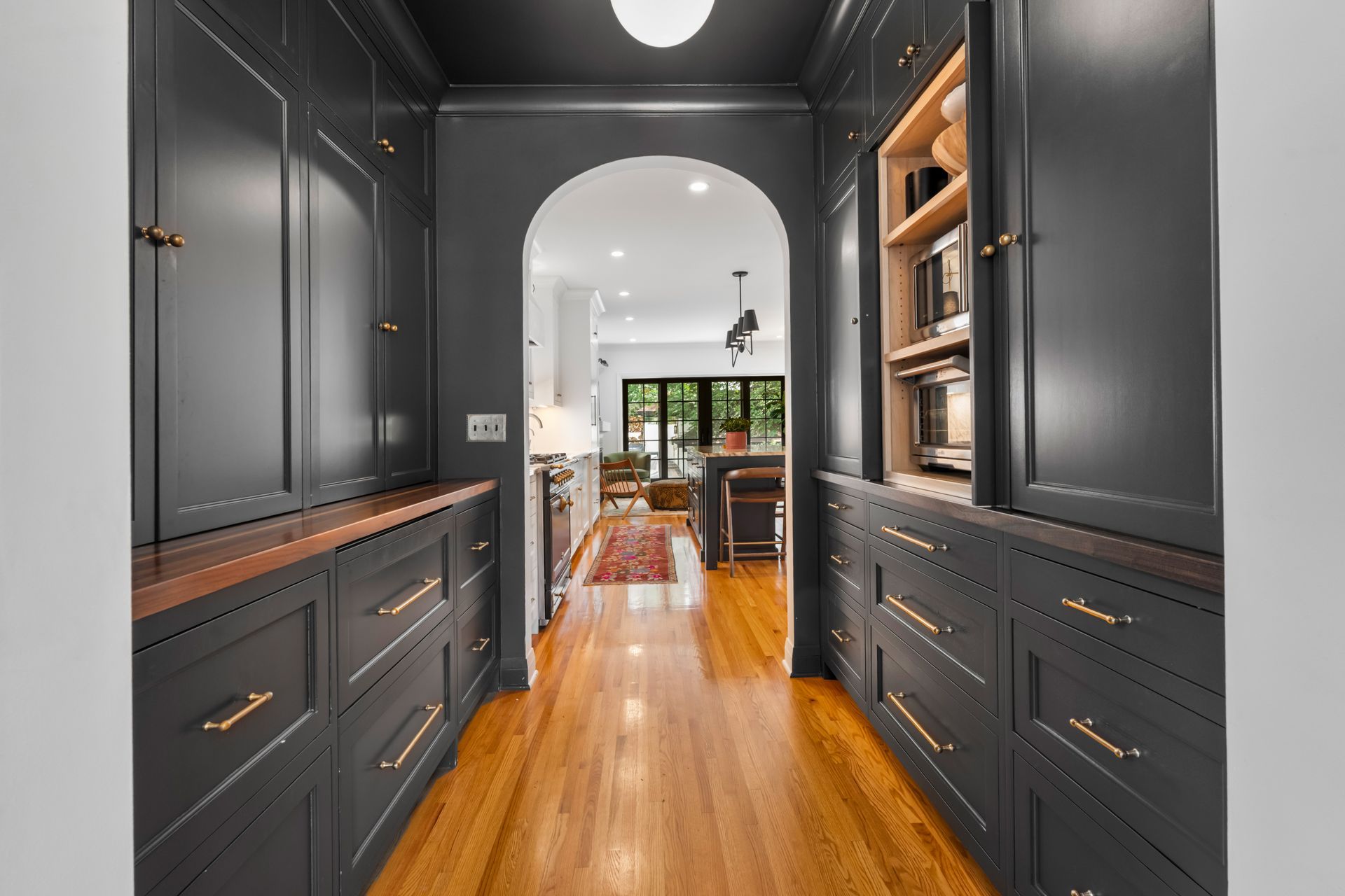 A hallway featuring dark gray cabinets with gold hardware on both sides, leading through an archway to a dining area.