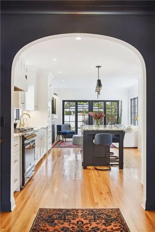 A view through an arched doorway into a bright kitchen and dining area featuring a dark island, white cabinets, and wood floors.