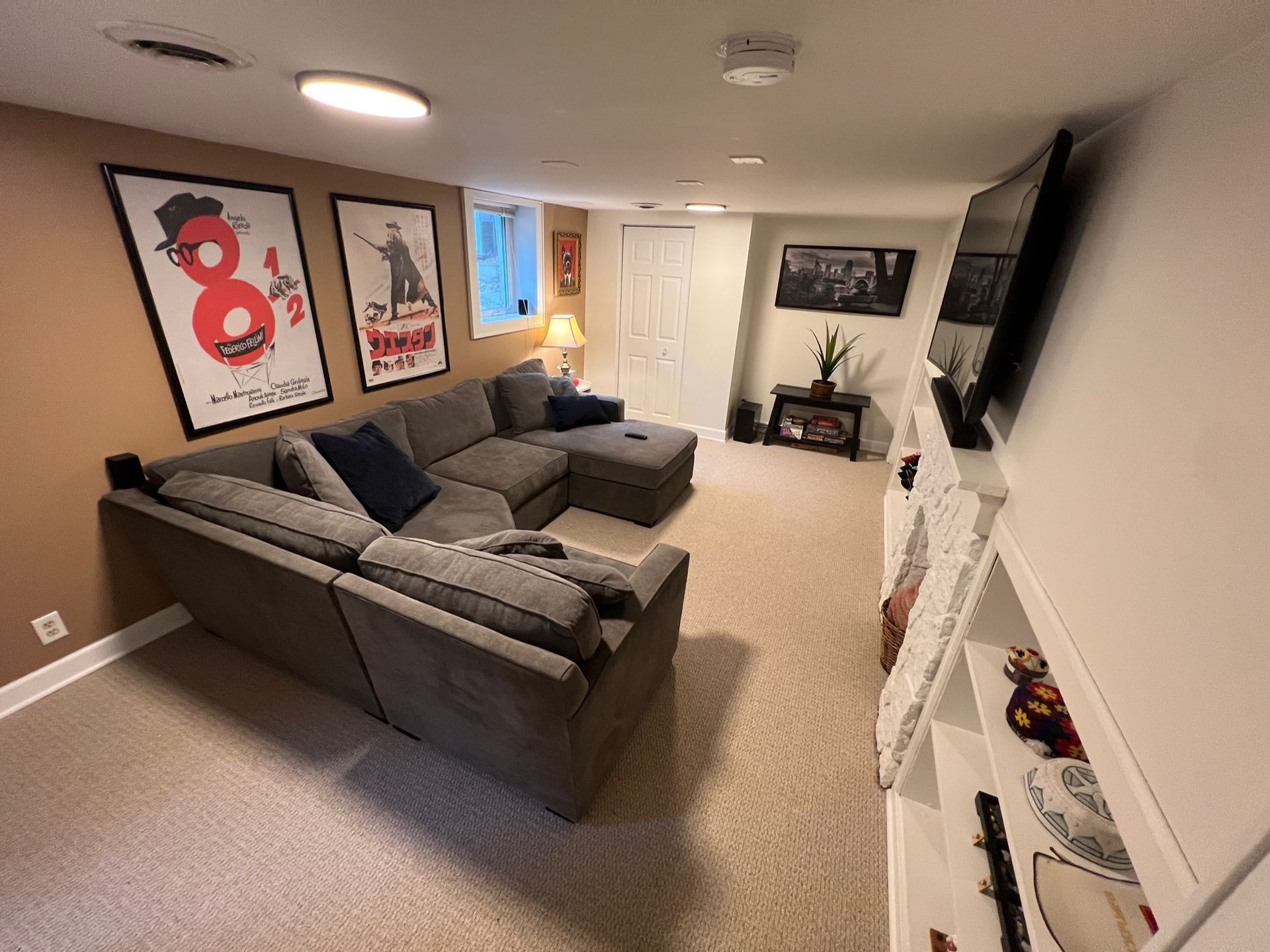 A basement living area with a large grey sectional sofa, two framed movie posters, carpet, a television, and shelving.
