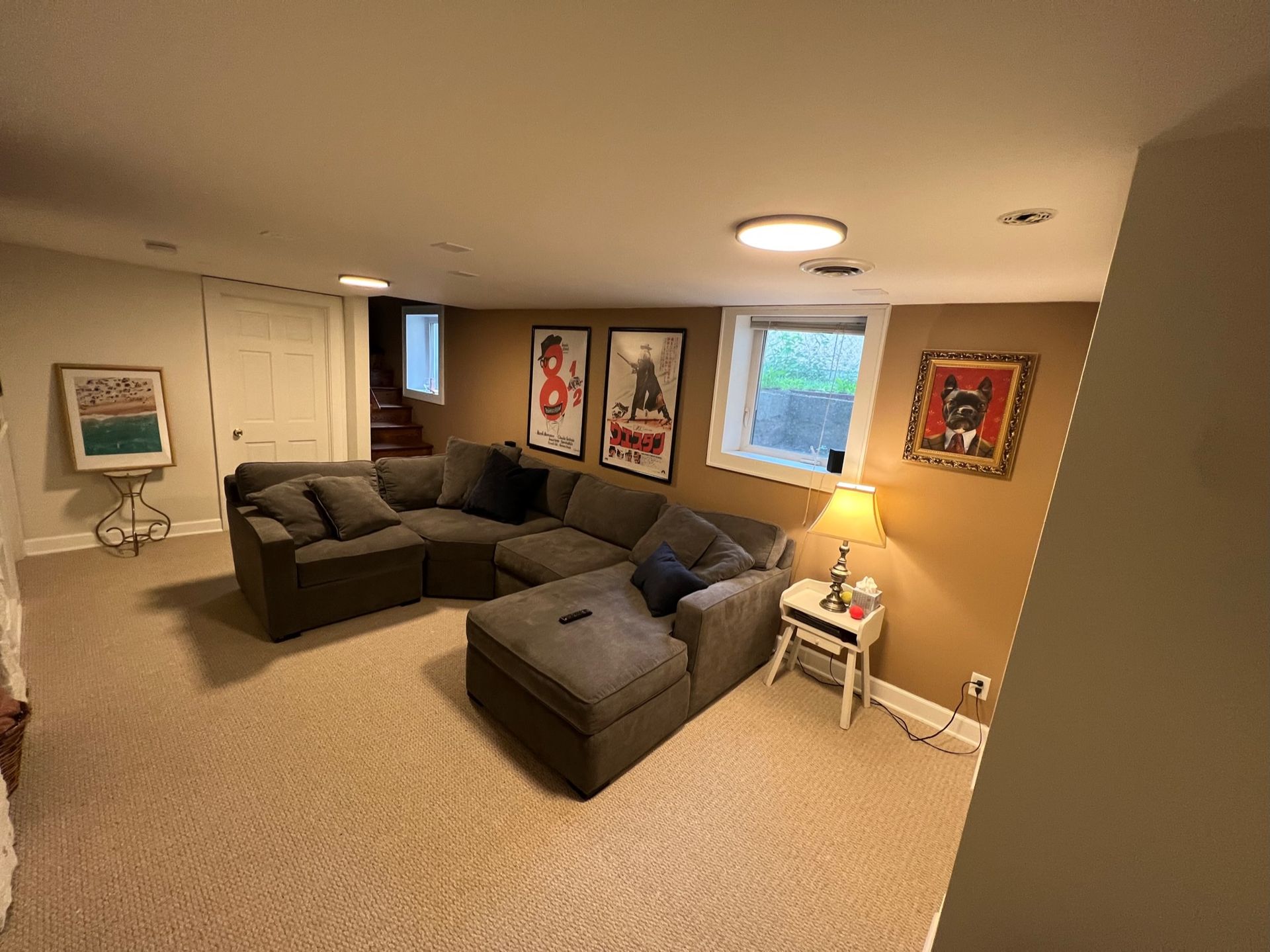 A cozy basement living room with a large gray sectional sofa, neutral carpet, framed art, and a small side table.