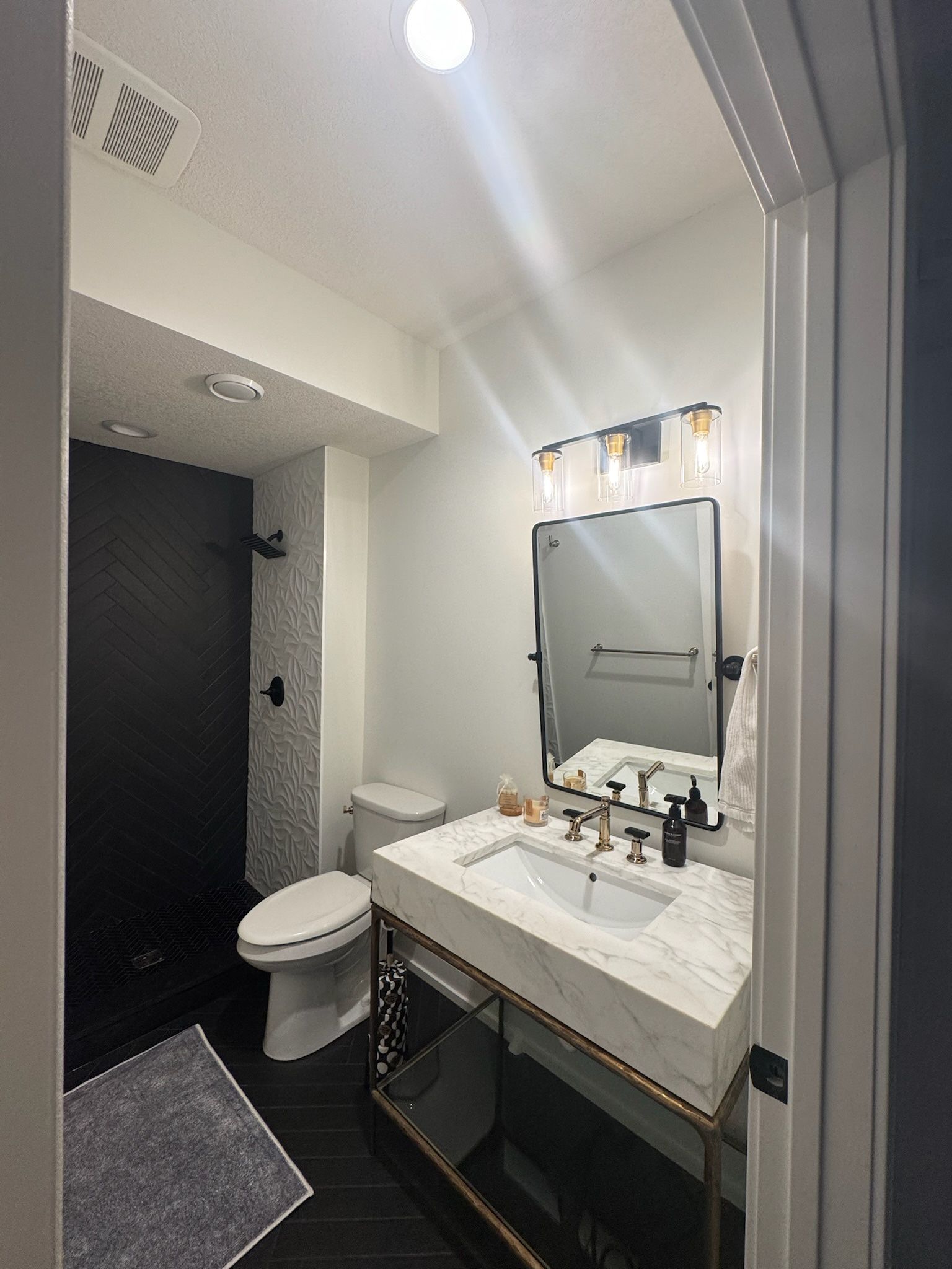 A modern bathroom with a vanity, marble countertop, mirror, light fixture, toilet, and a black-tiled shower.