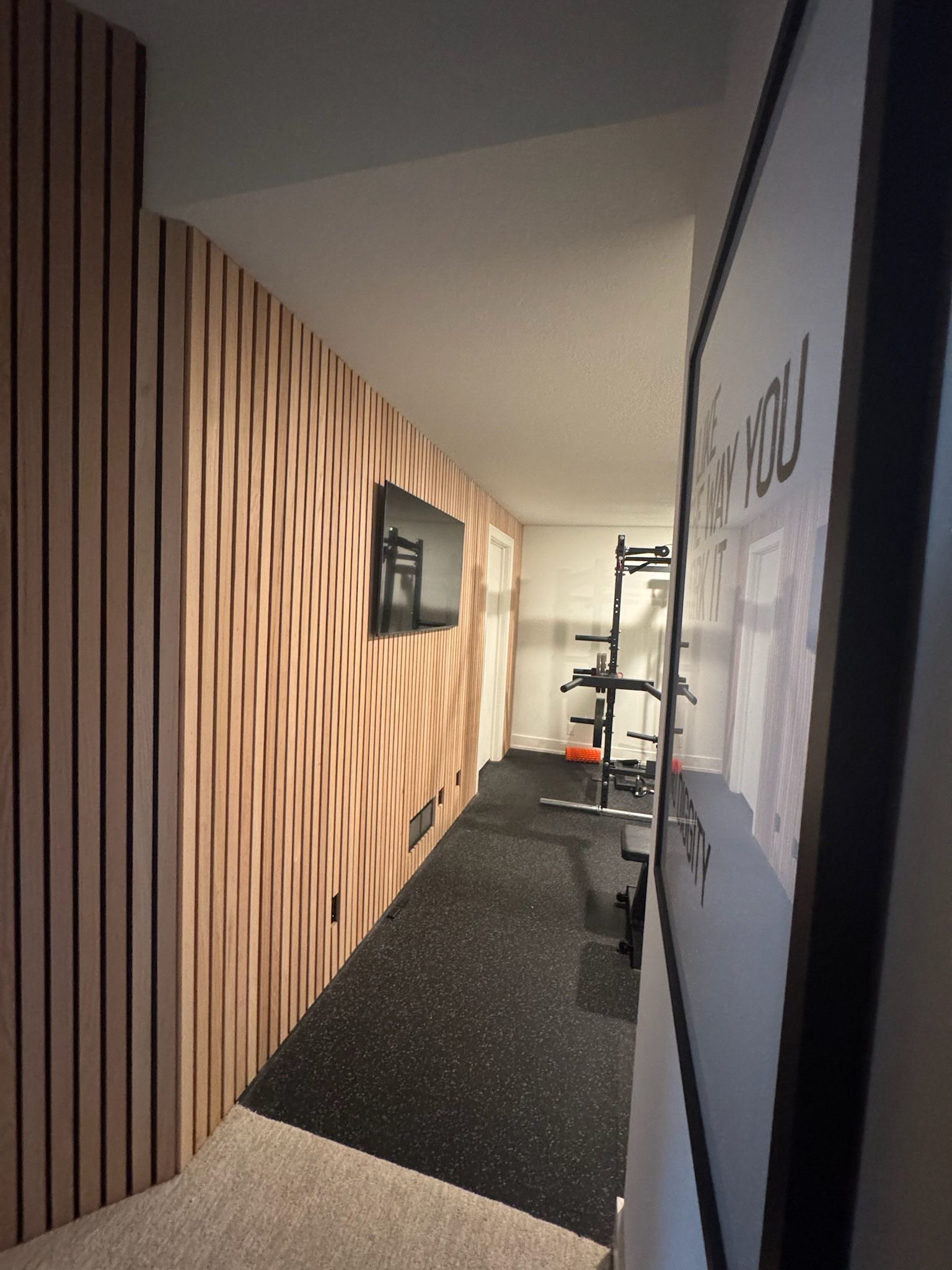 A hallway with a wood-slat feature wall, leading to a home gym with workout equipment and black rubber flooring.