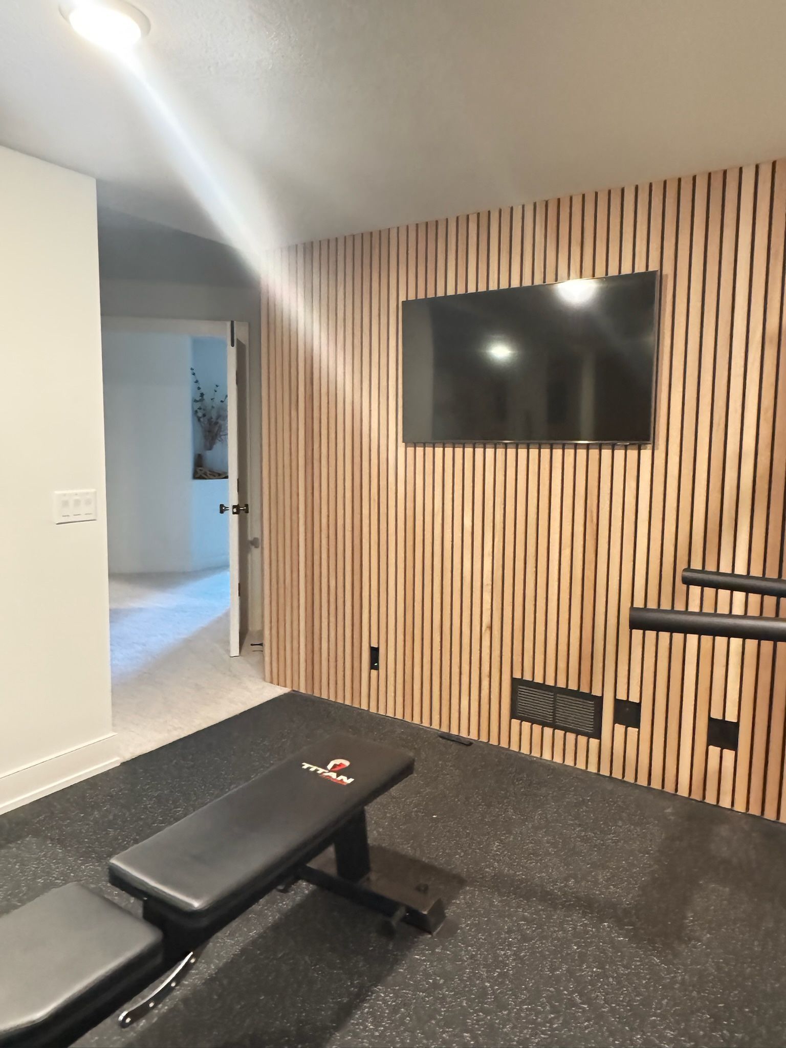 A home gym with a wall featuring vertical wood slats, a mounted television, and a workout bench on a black rubber floor.