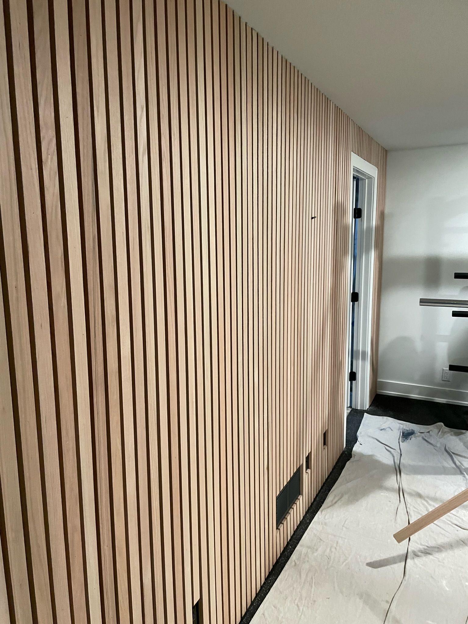 A wood-paneled wall featuring alternating wavy, vertical light wood planks and dark recessed gaps inside a room.