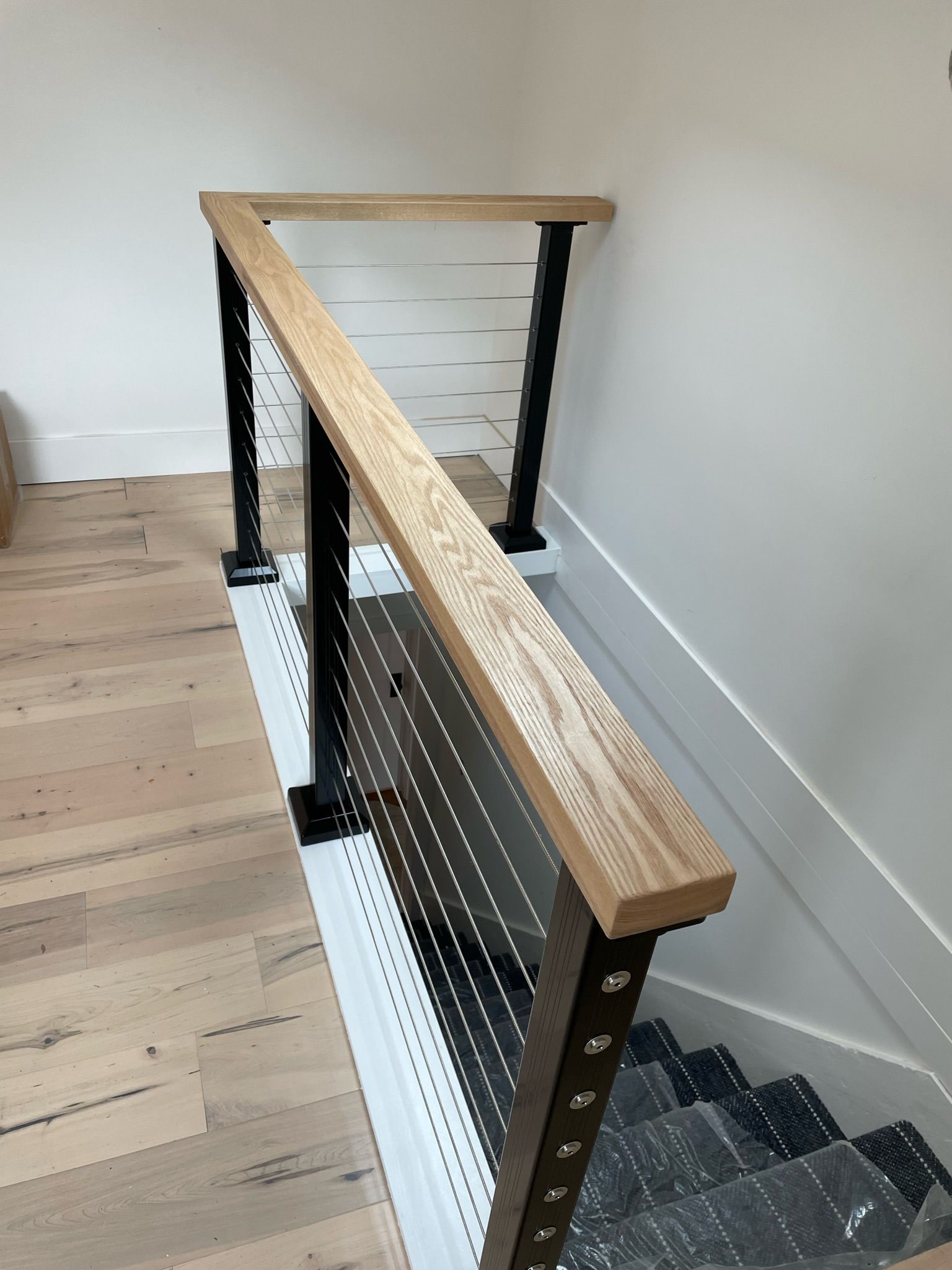 A wooden handrail atop black metal balusters and a white decorative panel overlooking a staircase with light wood flooring.