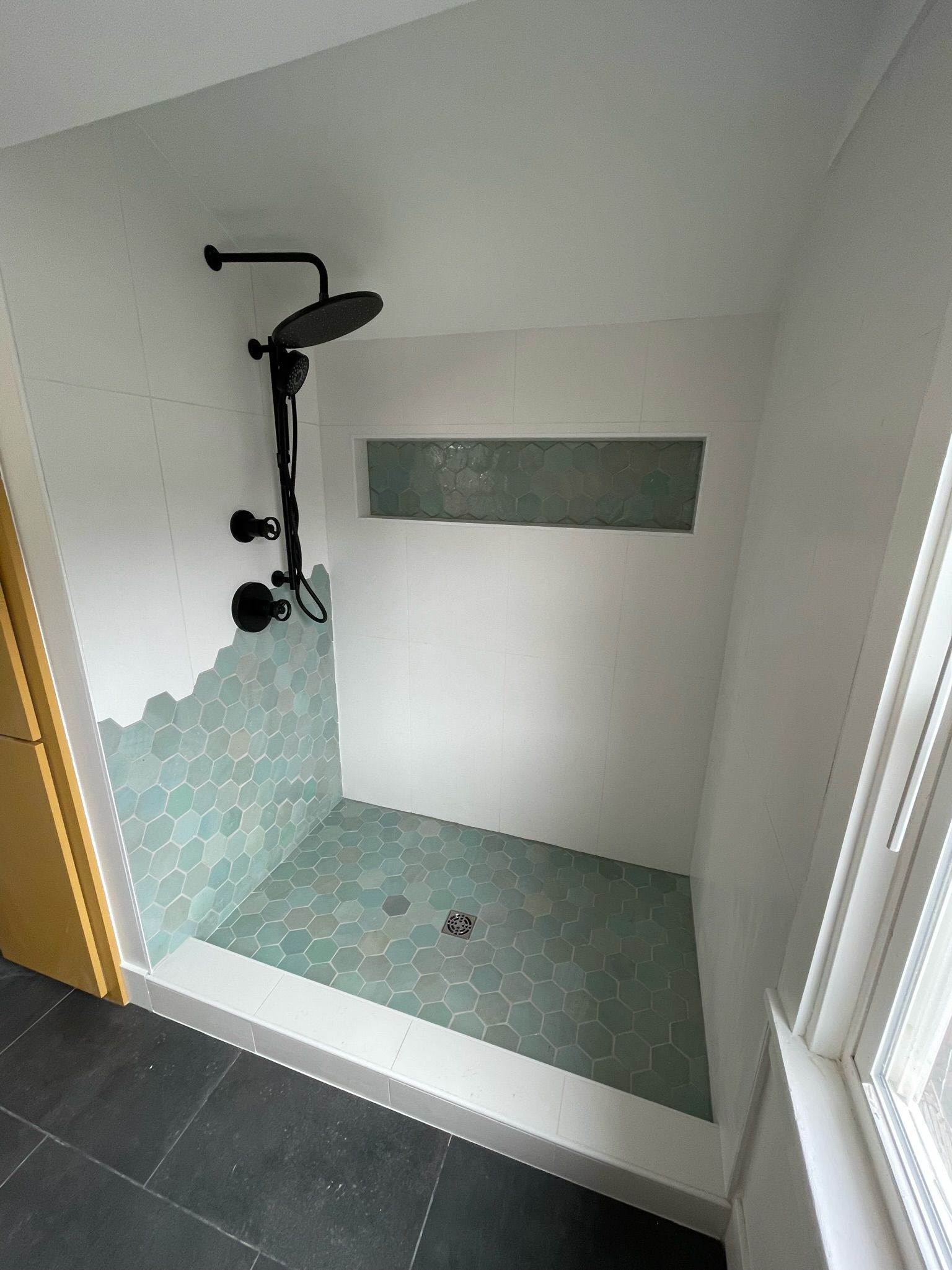 A partially tiled walk-in shower with green hexagonal tiles, a black rainfall shower head, and a recessed wall shelf.