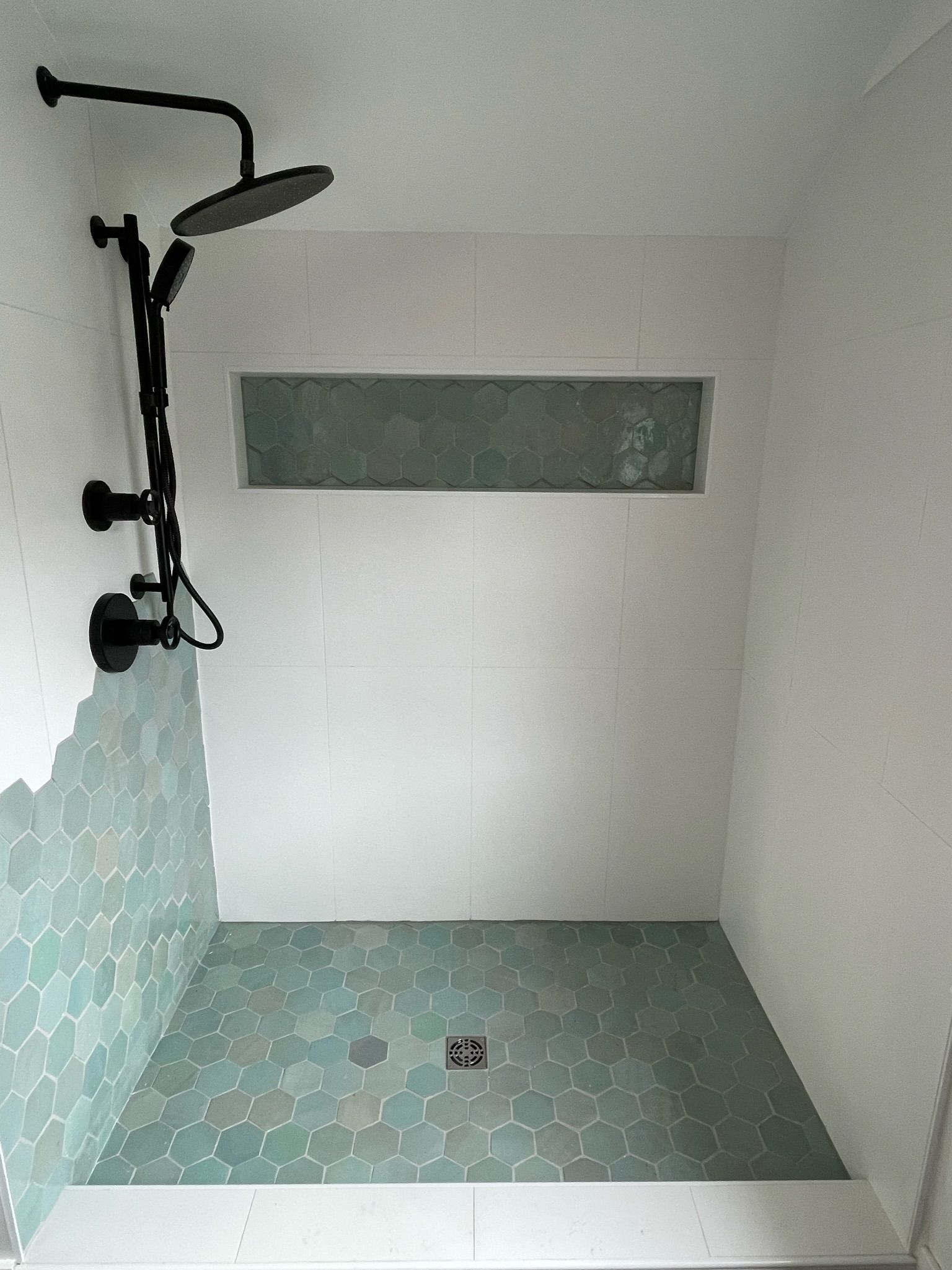 A modern walk-in shower with a black rainfall showerhead, white walls, and blue-green hexagonal tile on the floor and wall.