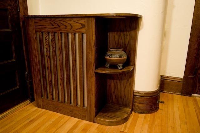 A custom wood radiator cover with an integrated display shelf, placed against a curved wall in a room with hardwood floors.