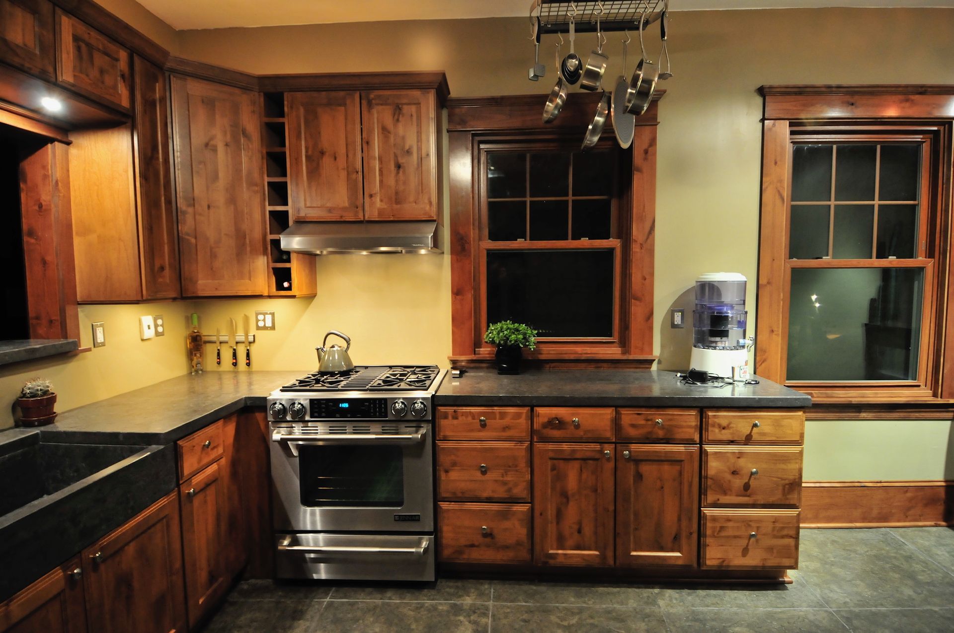 A rustic-style kitchen with natural wood cabinets, dark stone countertops, stainless steel appliances, and two windows.
