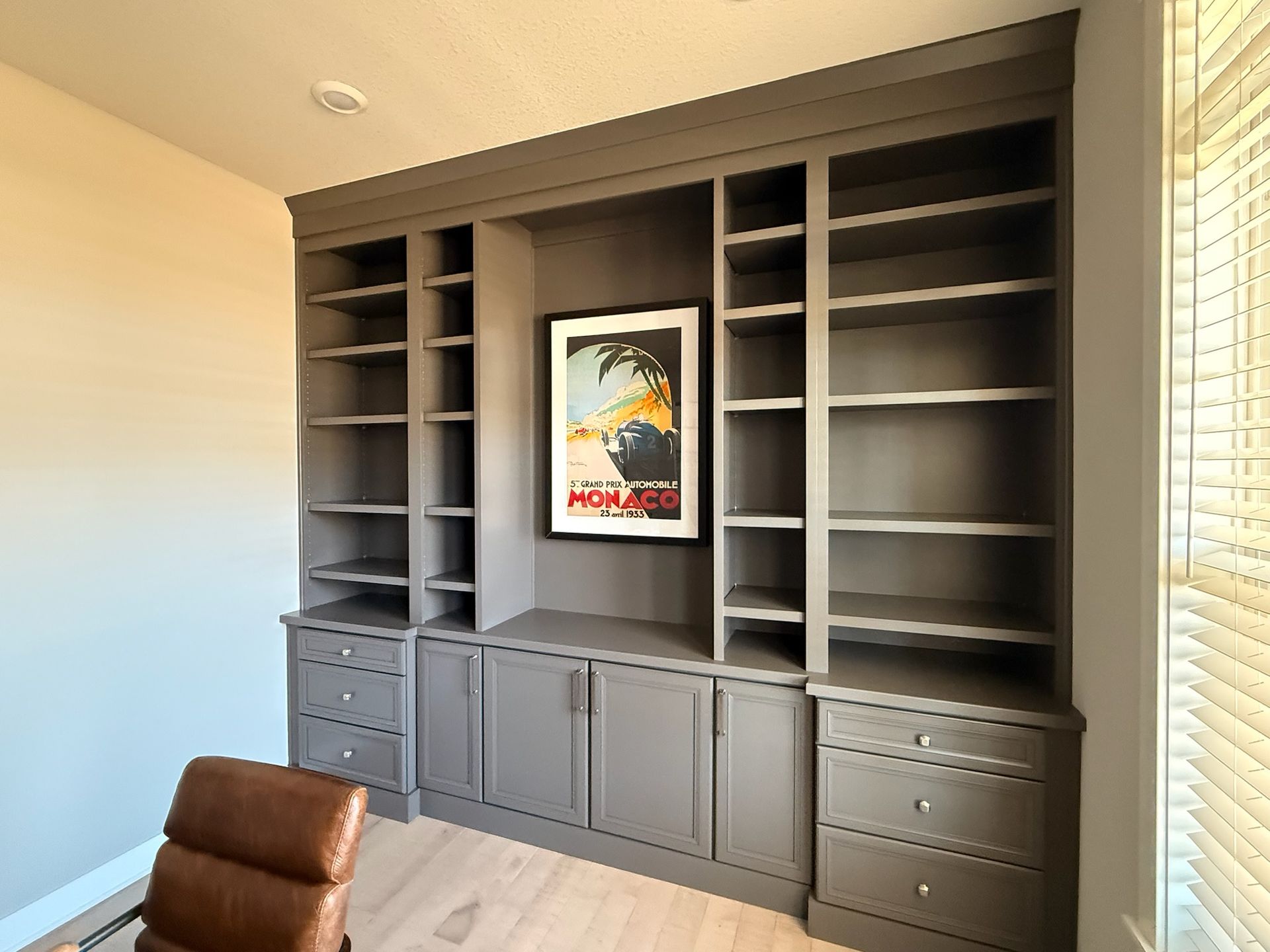 Built-in grey shelving unit with open shelves, lower cabinets, and drawers flanking a framed Monaco travel poster.