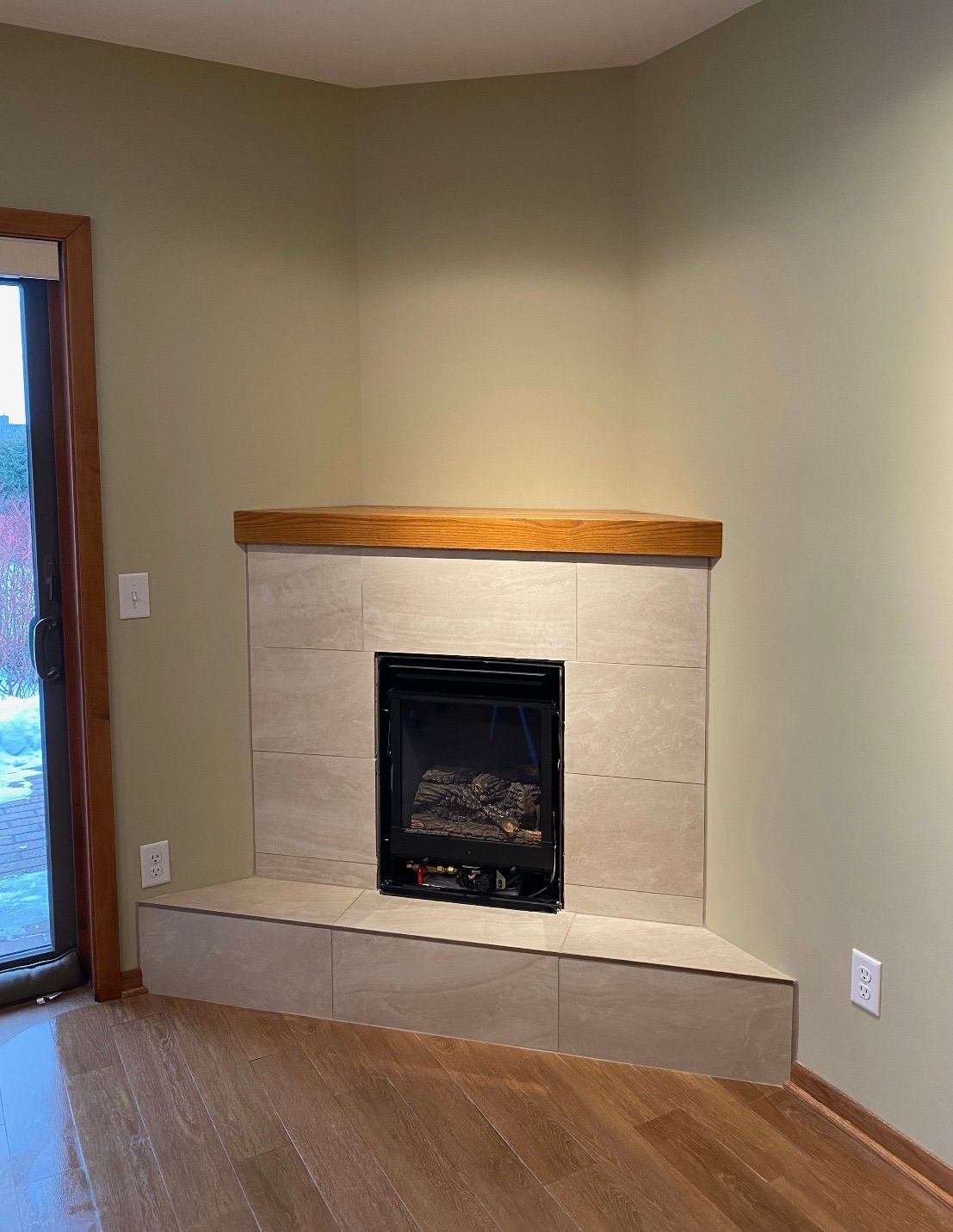 A corner fireplace with a dark insert, neutral-toned square tile surround and hearth, and a stained wood mantel.