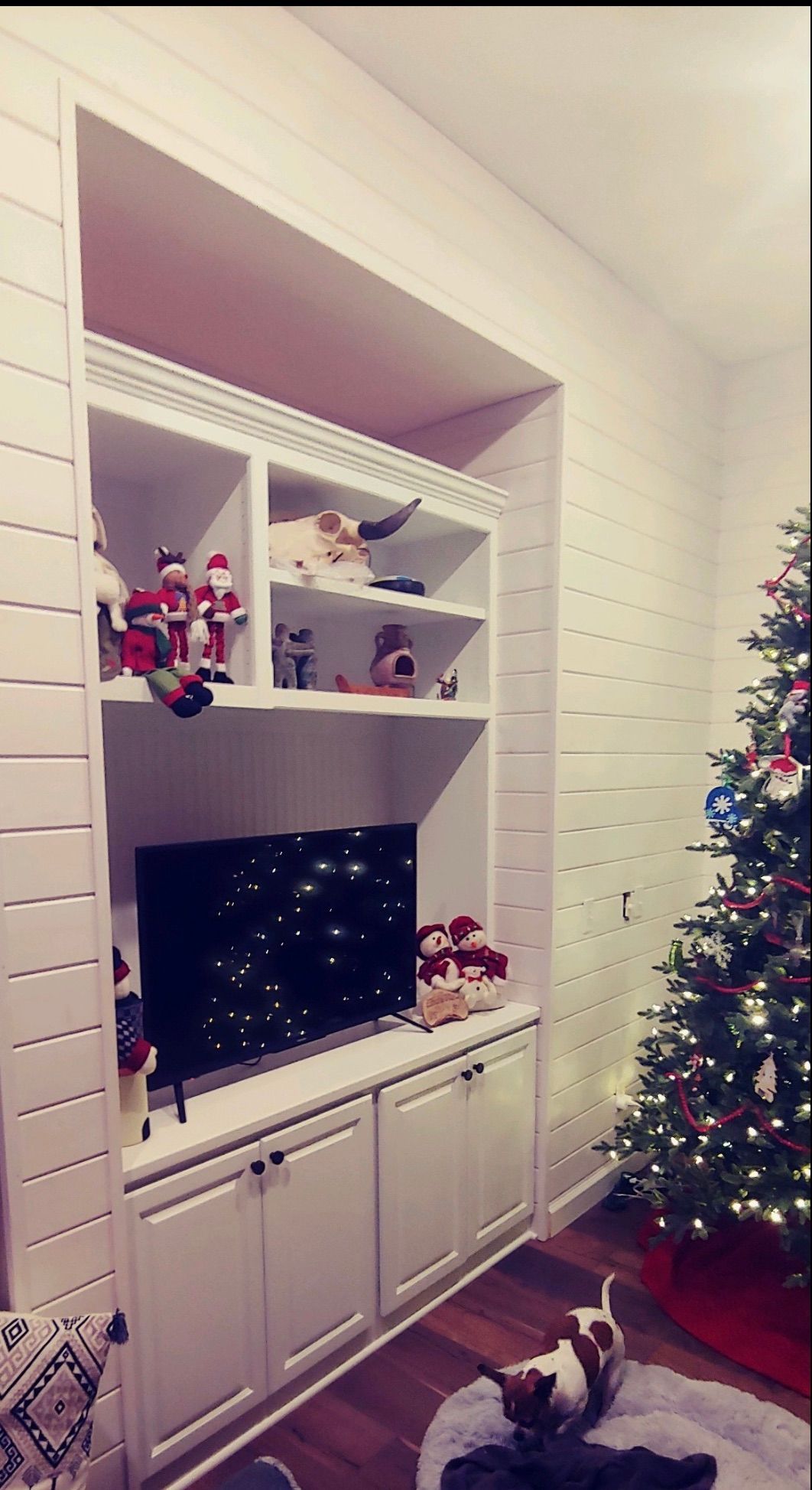 A white built-in shelf unit with holiday decor, a TV, and a dog lying on a nearby rug next to a decorated Christmas tree.