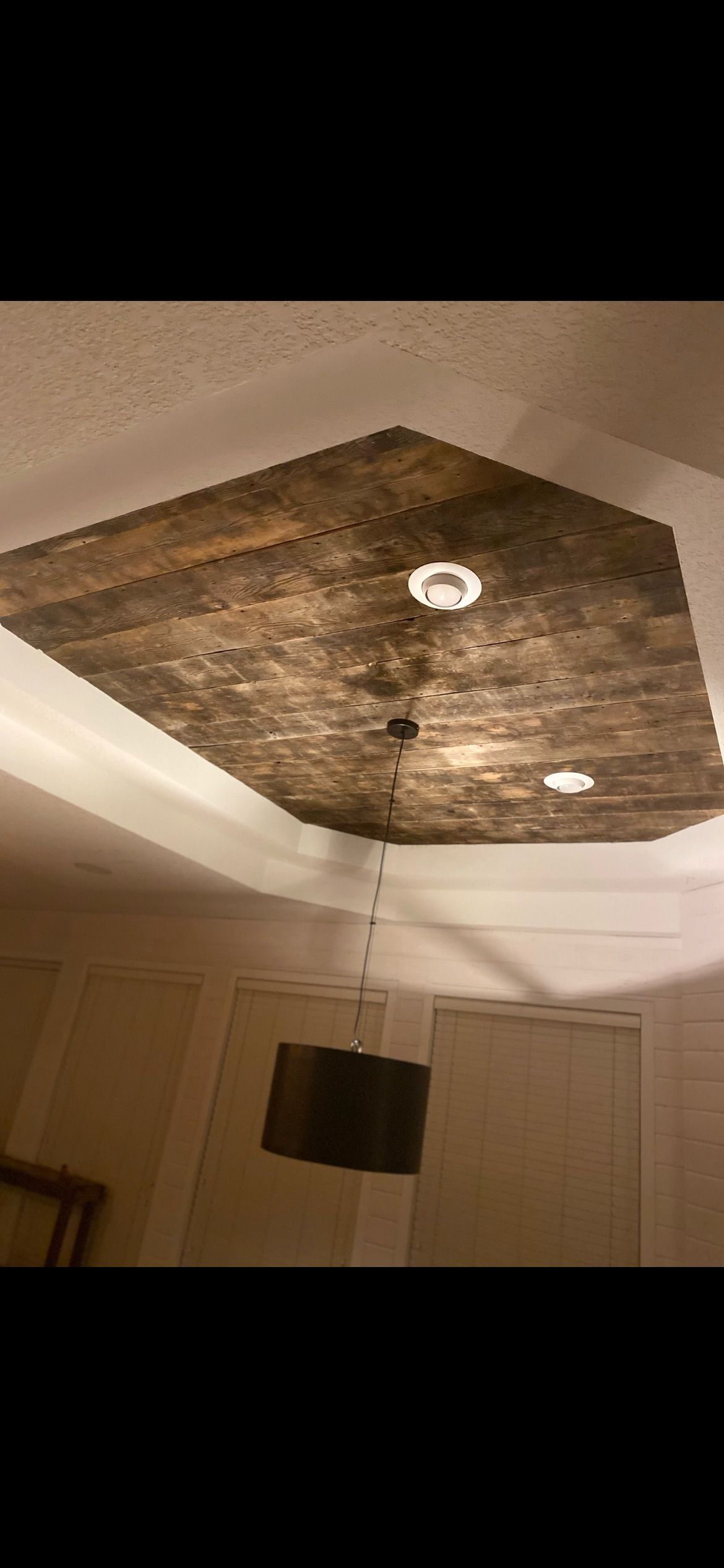 A low-angle view of a tray ceiling featuring dark, textured wood planks and recessed lighting above a hanging pendant lamp.