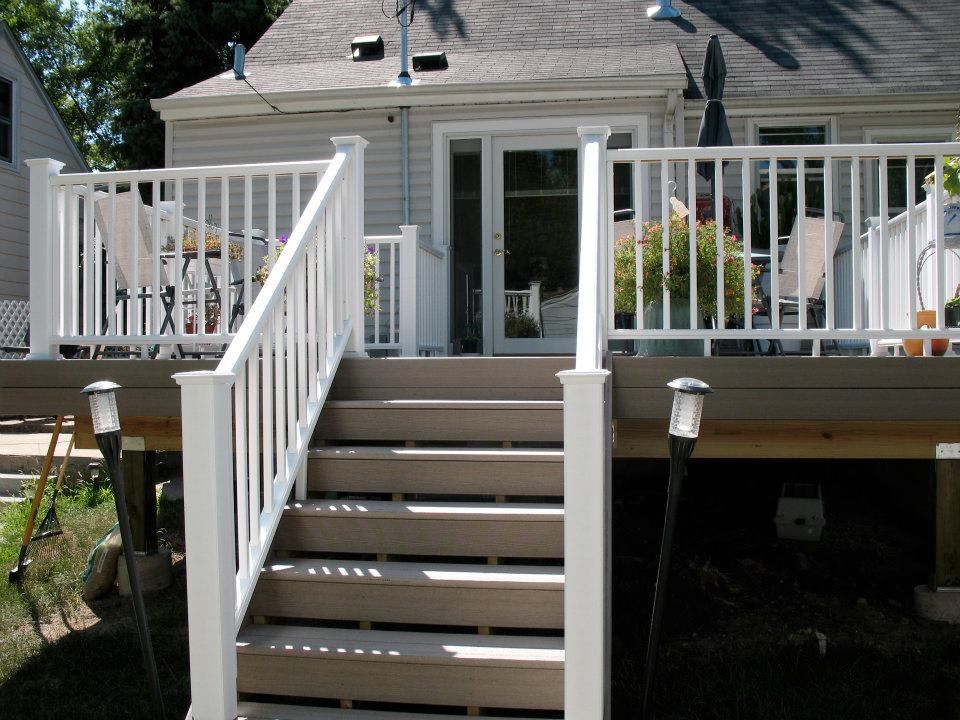A view of a tan composite deck with white railings and stairs leading to a back door of a house with grey siding.