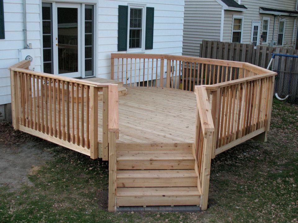 A light wood deck with railings and steps leading to the backyard of a white house.