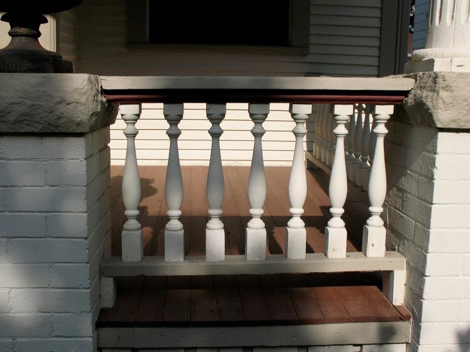 White wooden balusters forming a porch railing between two brick pillars on a porch with brown wooden decking.