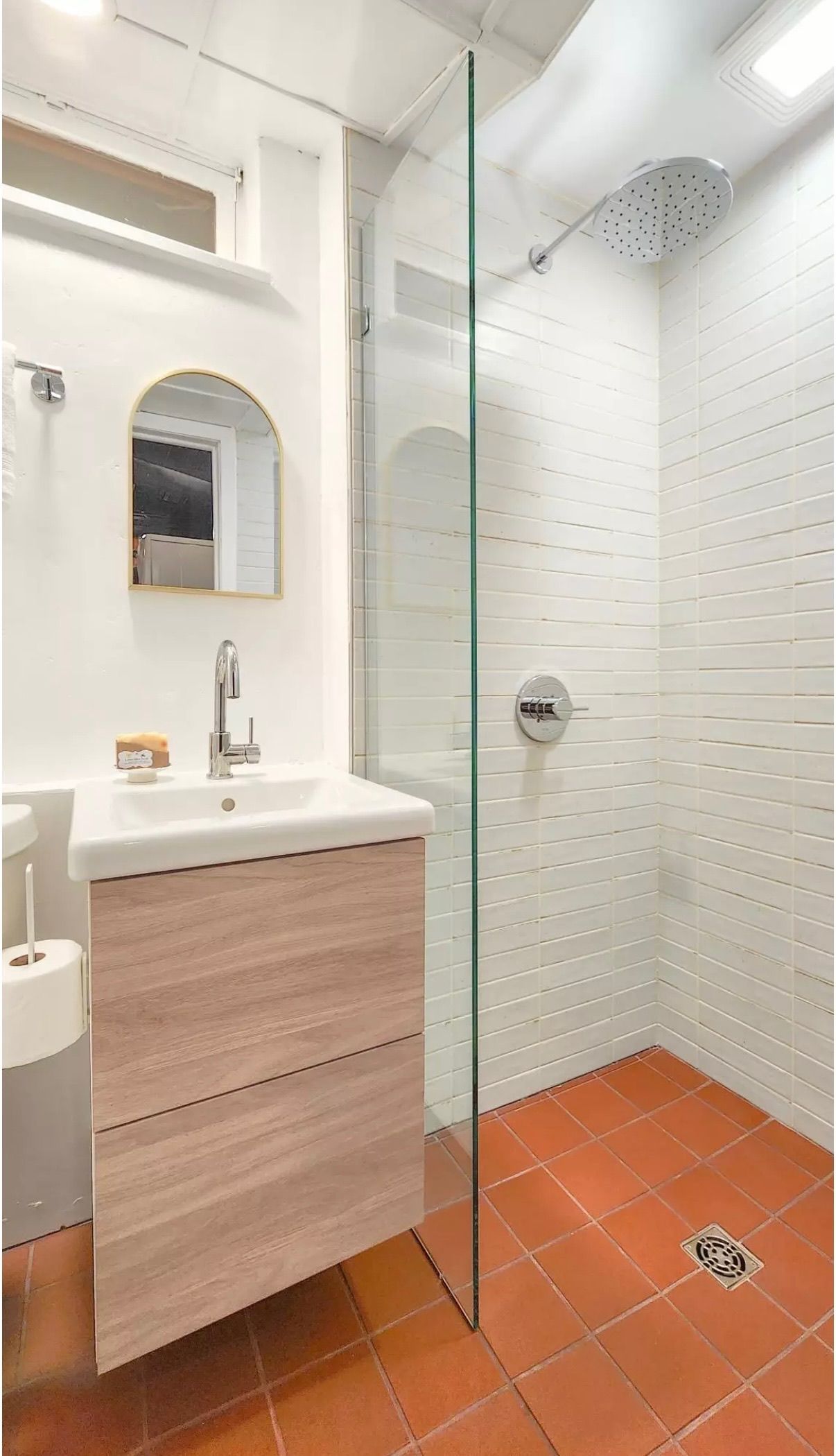 A bathroom with a floating light-wood vanity, an arched mirror, and a walk-in glass shower with terracotta floor tiles.