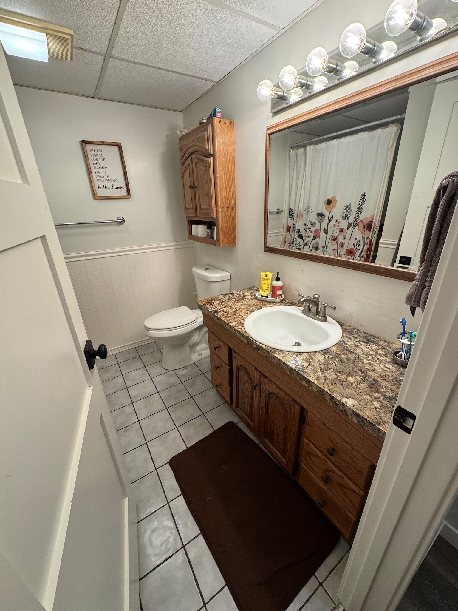 A small bathroom with a wood vanity, brown countertop, beige walls, white tile floor, and a floral shower curtain.