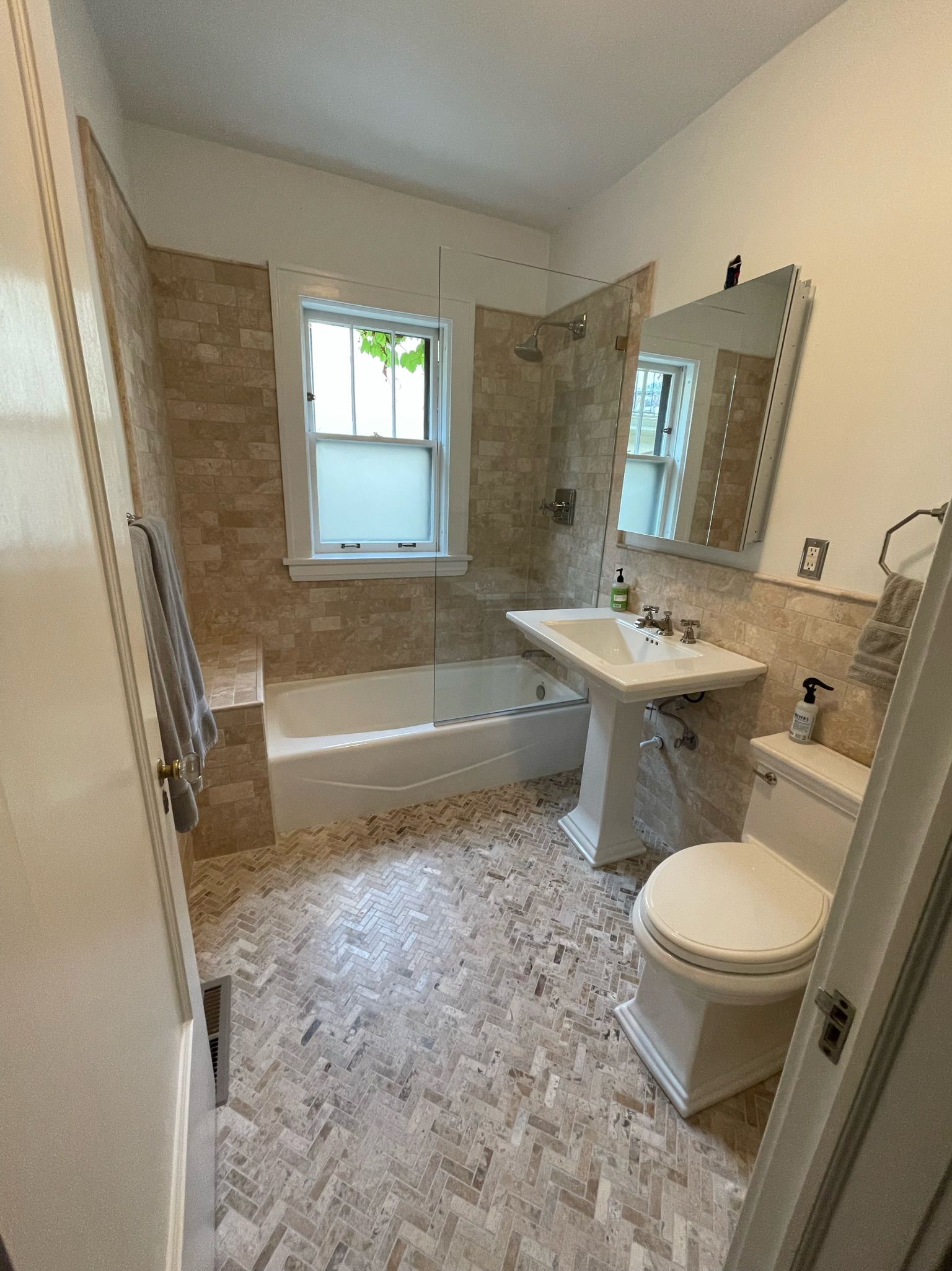 A bright bathroom with beige tile walls and flooring, featuring a bathtub with a window, pedestal sink, and white toilet.