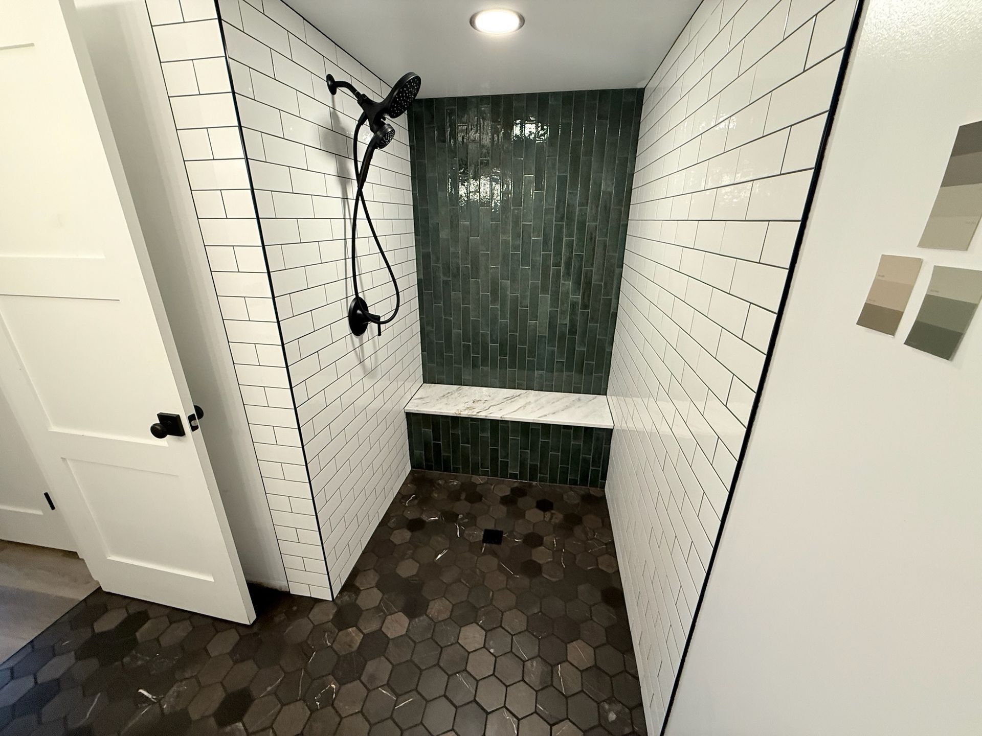 A walk-in shower with white subway tile walls, a dark green tiled back wall, a marble bench, and dark hexagonal floor tiles.