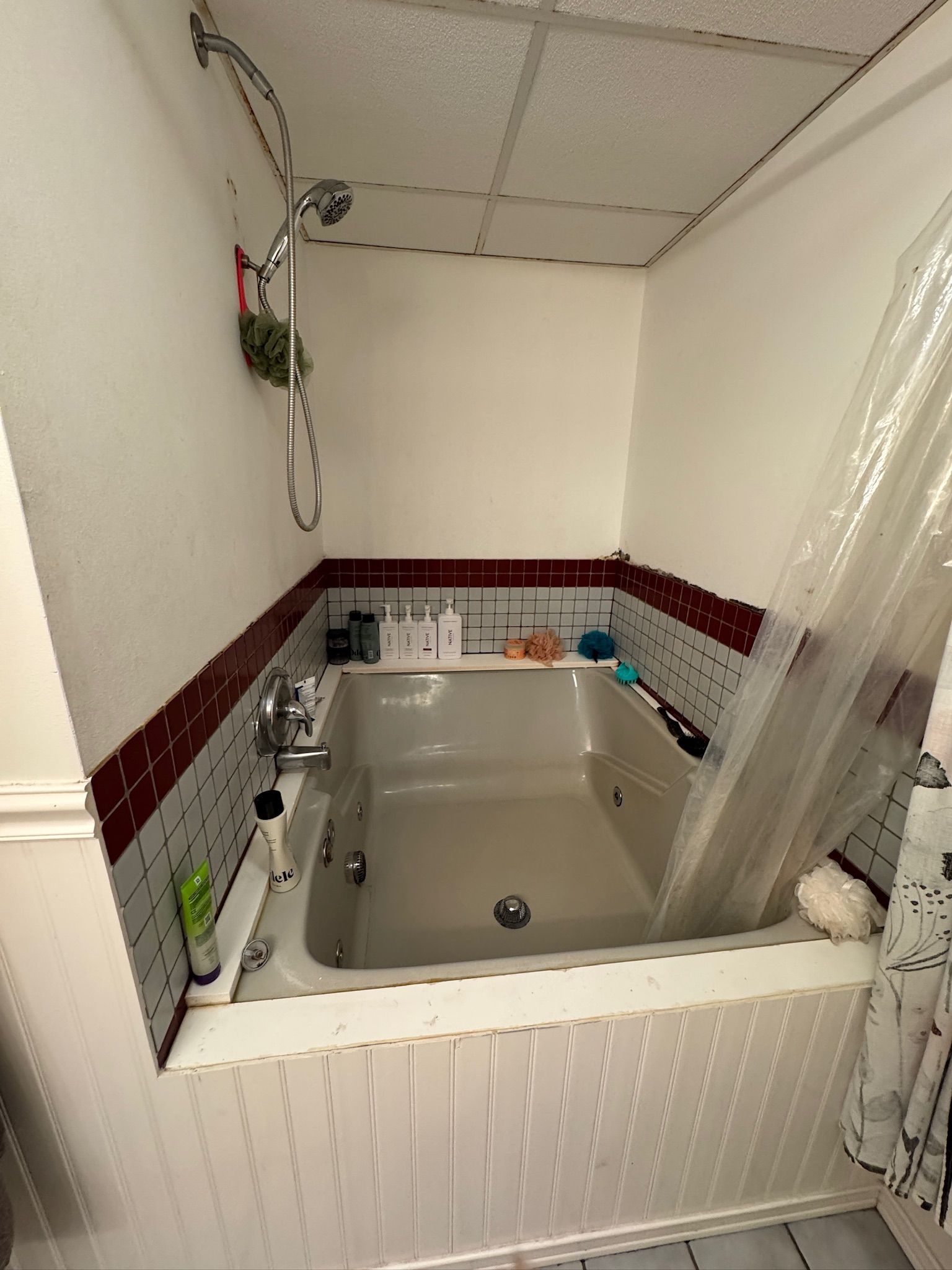 A built-in bathtub with a shower head, surrounded by dark red and grey tiled walls and white vertical paneling.