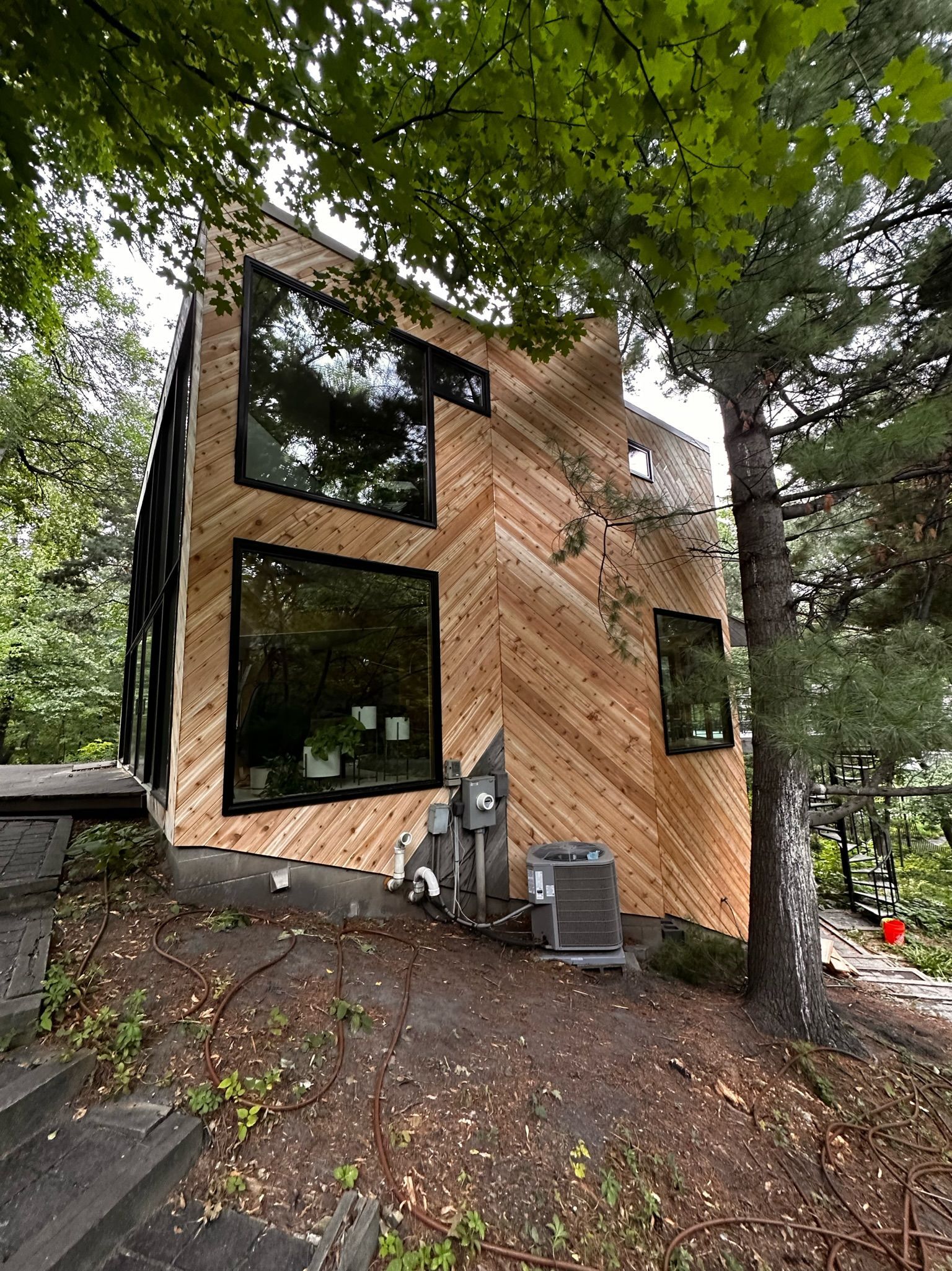 A two-story house featuring natural wood siding in a herringbone pattern, large square windows, and a shaded yard.