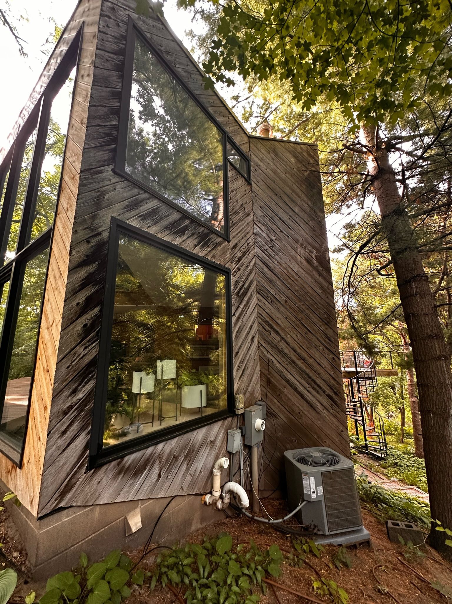 Modern cabin with diagonal wood siding and large windows, surrounded by trees on a wooded slope with an AC unit nearby.