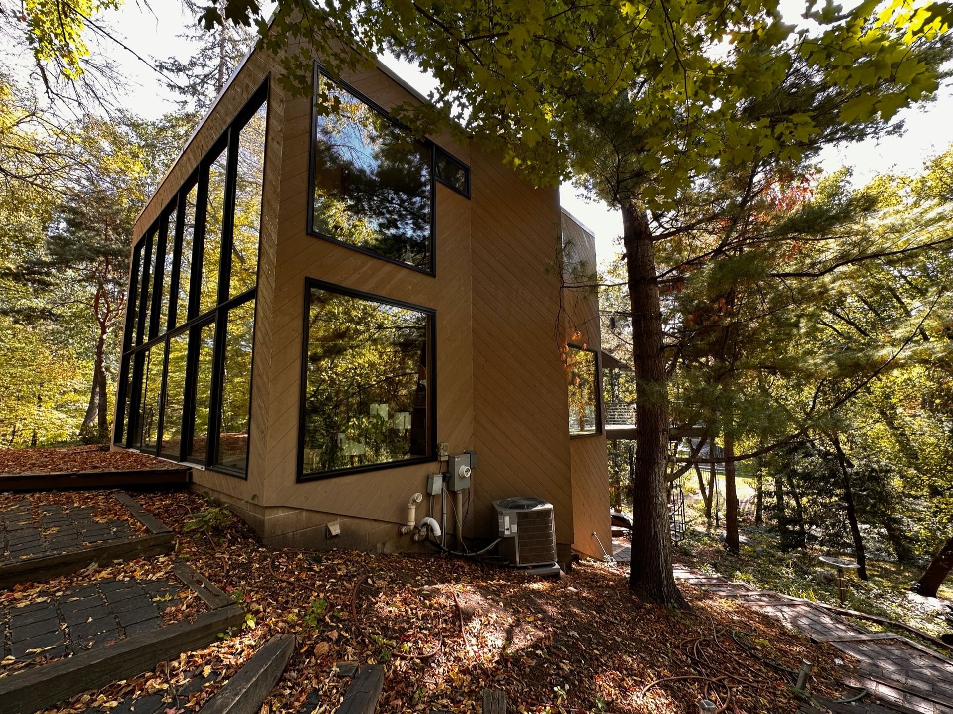 A modern house with large windows and tan exterior walls nestled in a wooded, leafy environment during autumn.