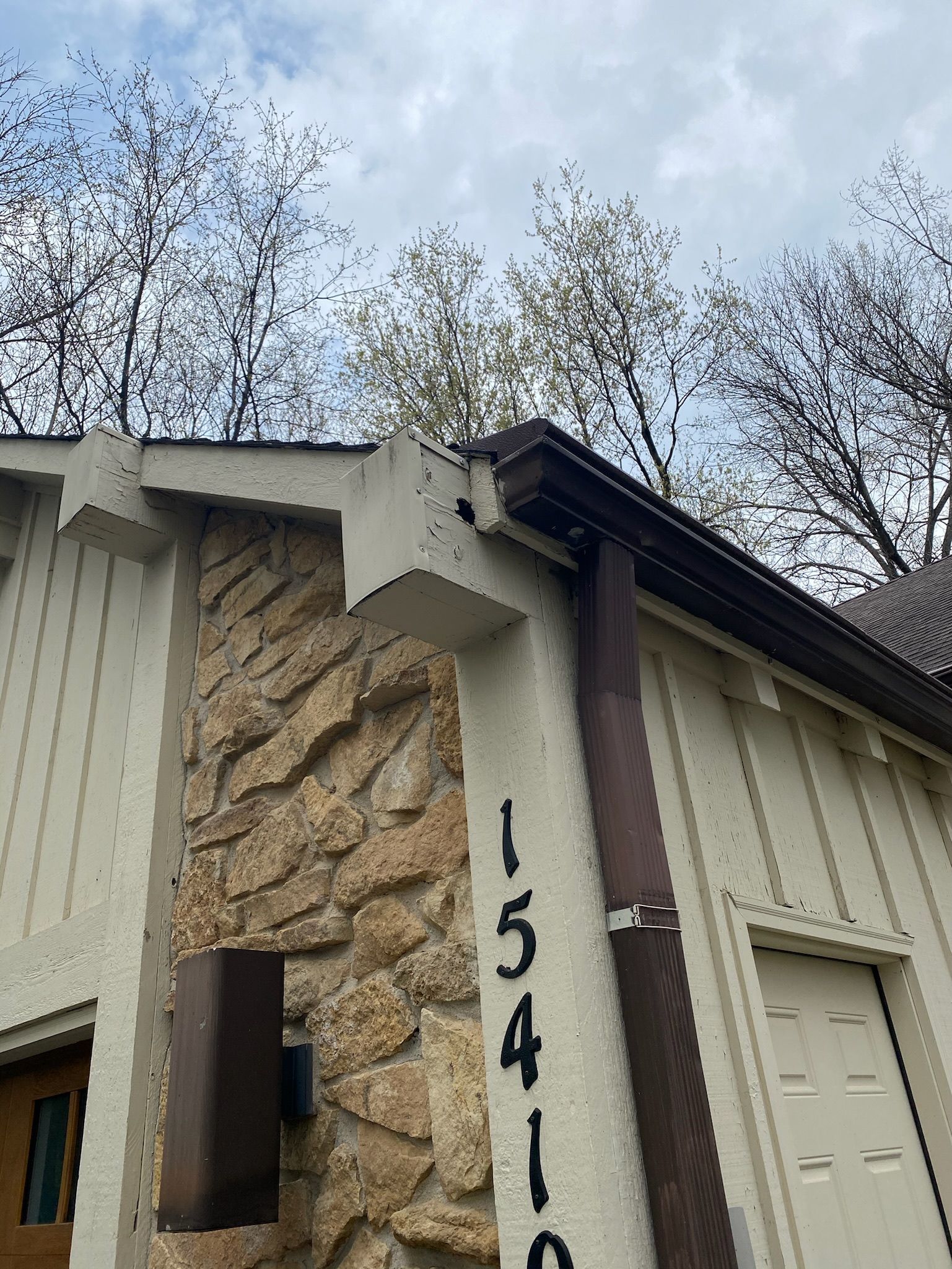 A low-angle view of a building corner with vertical siding, stone masonry, a dark downspout, and house number 15410.