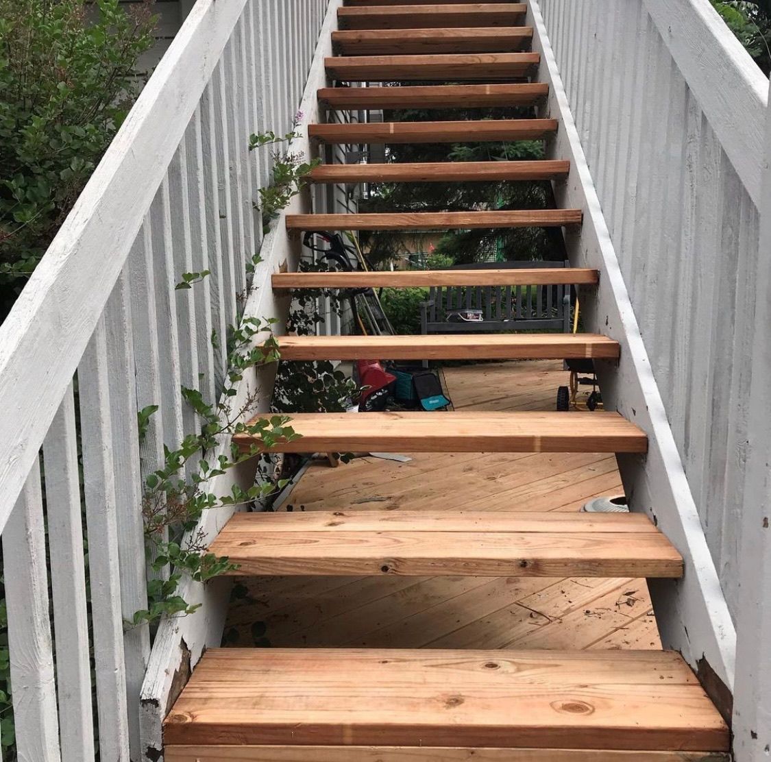 A set of wooden stairs with white railings outdoors, leading up toward trees and a shaded deck area.