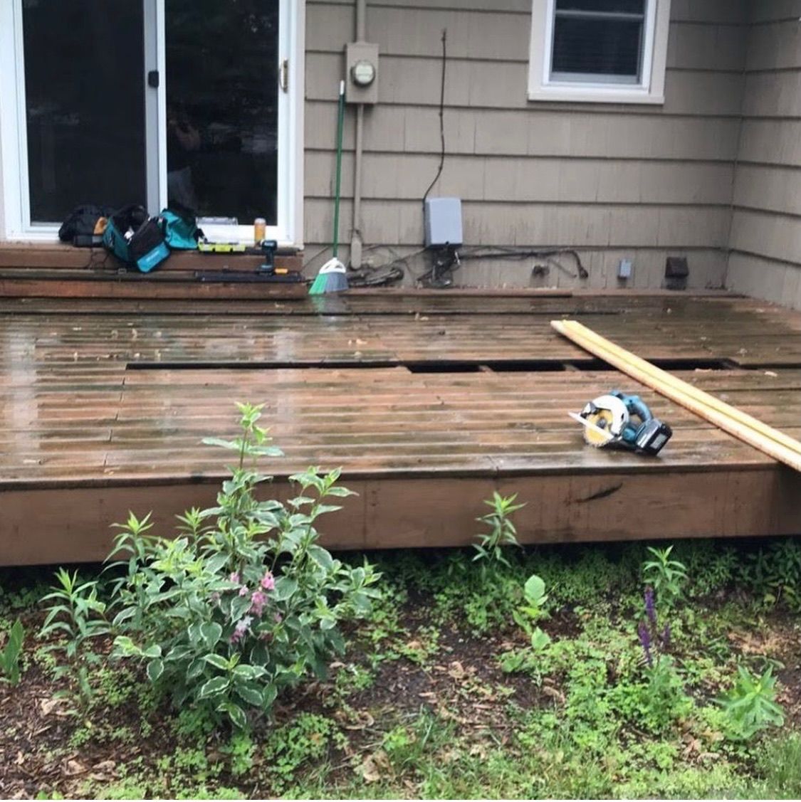 A residential wooden deck under repair, featuring a missing plank, a circular saw, and wood boards resting on the surface.