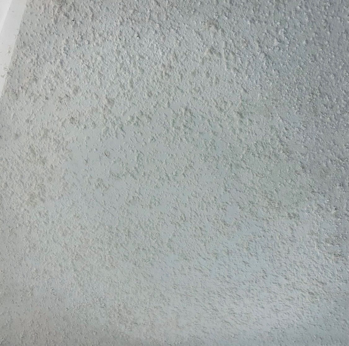 A close-up of a light gray wall with a rough, textured stucco or popcorn finish.