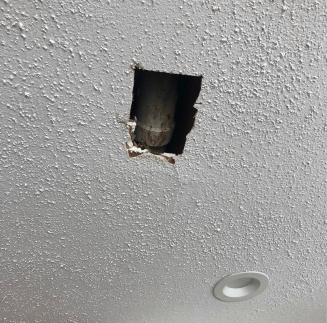 A hole in a textured white ceiling exposing a vertical pipe, with a recessed light fixture visible nearby.